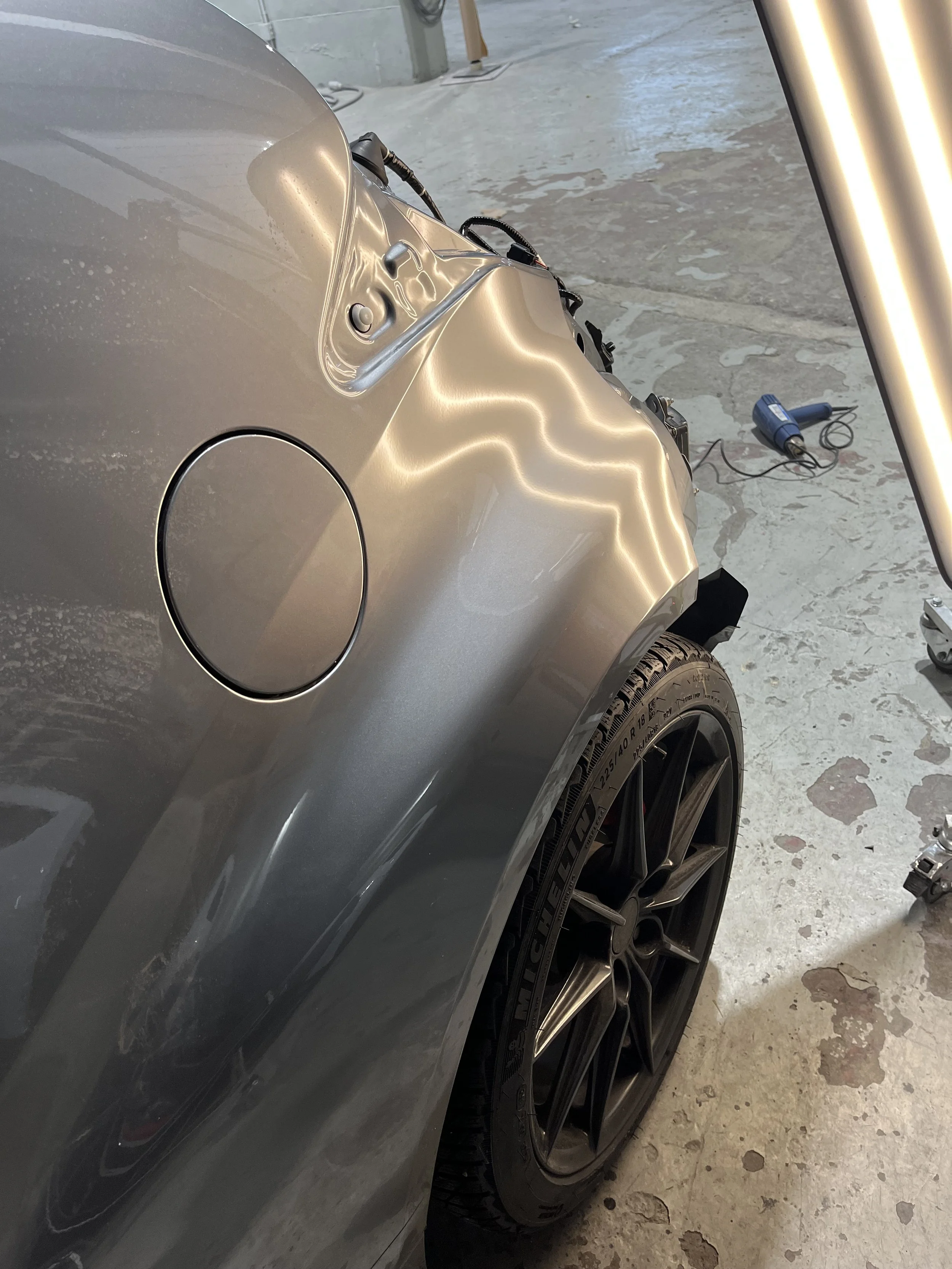 Close-up of a gray sports car in a garage or workshop with a black wheel and tire, electric drill, and garage flooring visible.