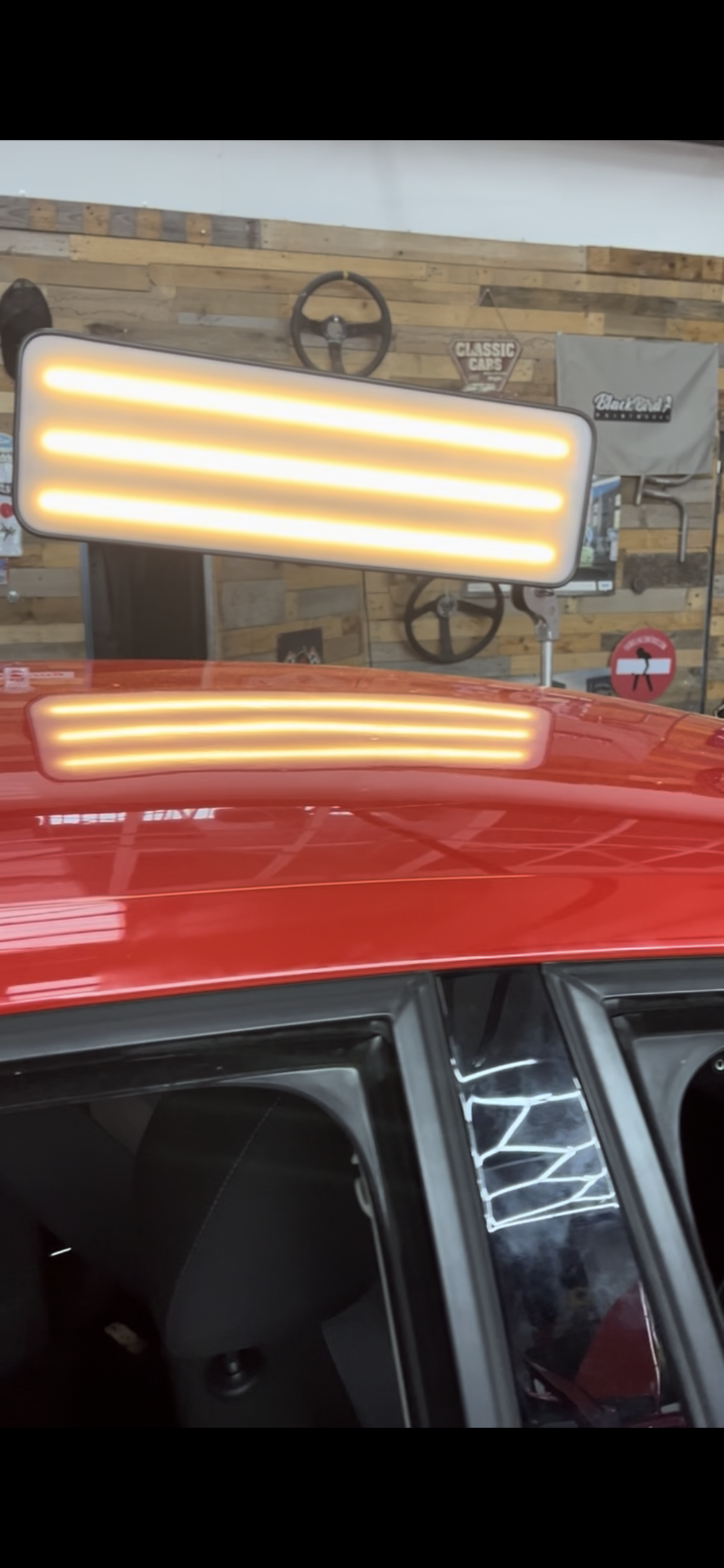 A red vehicle with illuminated yellow signal lights on top, reflected in the vehicle's roof. The background features a wooden wall with vintage car and road signs, gauges, and automotive memorabilia.
