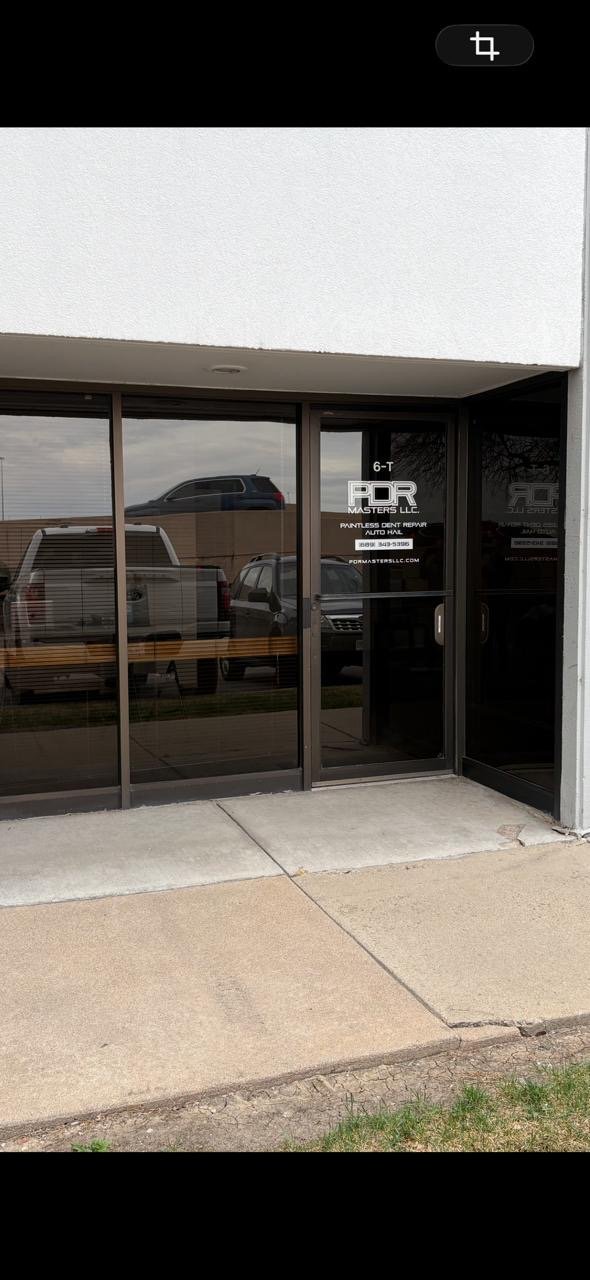 The storefront has glass doors and windows with reflections of vehicles parked outside. There is a sign on the door that reads "6-T PR Masters LLC" and advertising services for dent repair.