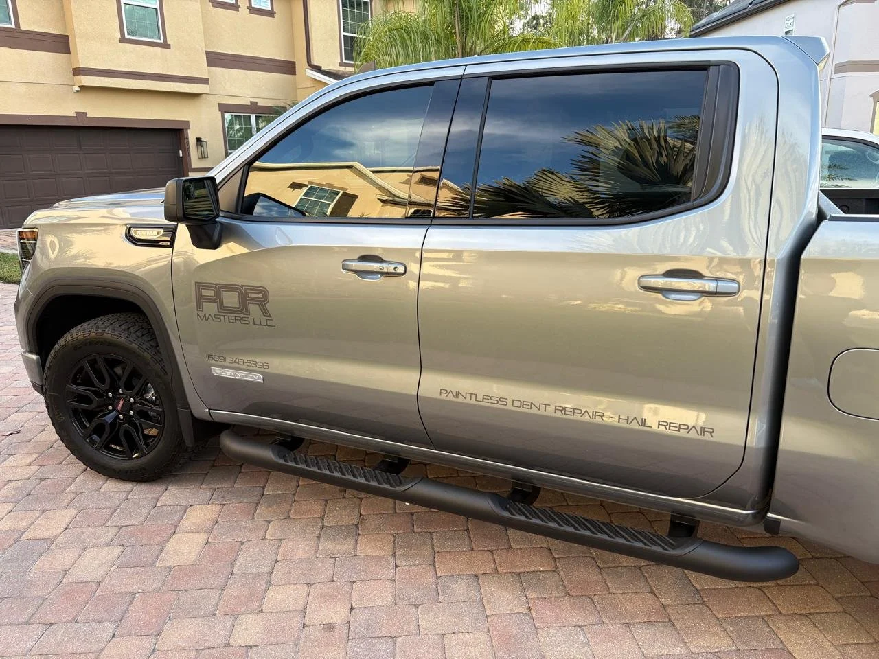 A silver pickup truck parked on a brick driveway, with black wheels, tinted windows, and decals that say 'PDR Masters LLC' and 'Paintless Dent Repair - Hail Repair,' in front of a residential building with palm trees.