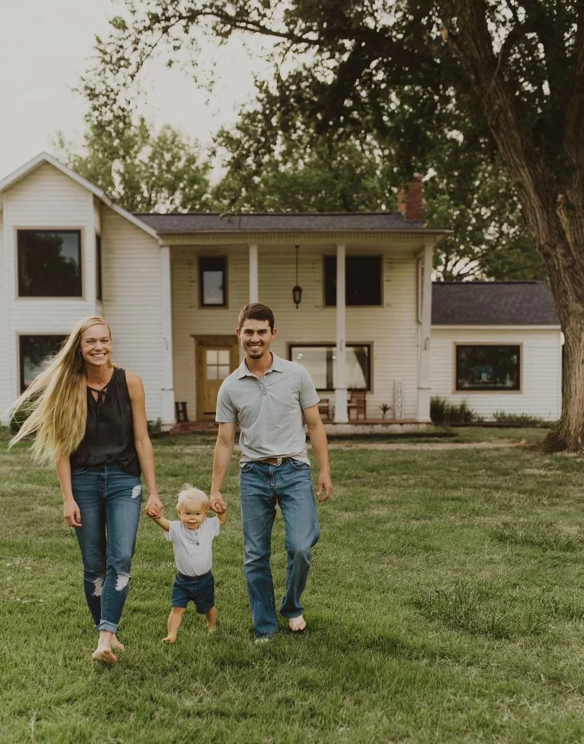 A family of three, including a woman, a man, and a young child, smiling and walking on a grassy lawn in front of a large two-story house with a porch, trees, and a clear sky in the background. They're grateful for Cornerstone Roofing & Exteriors.