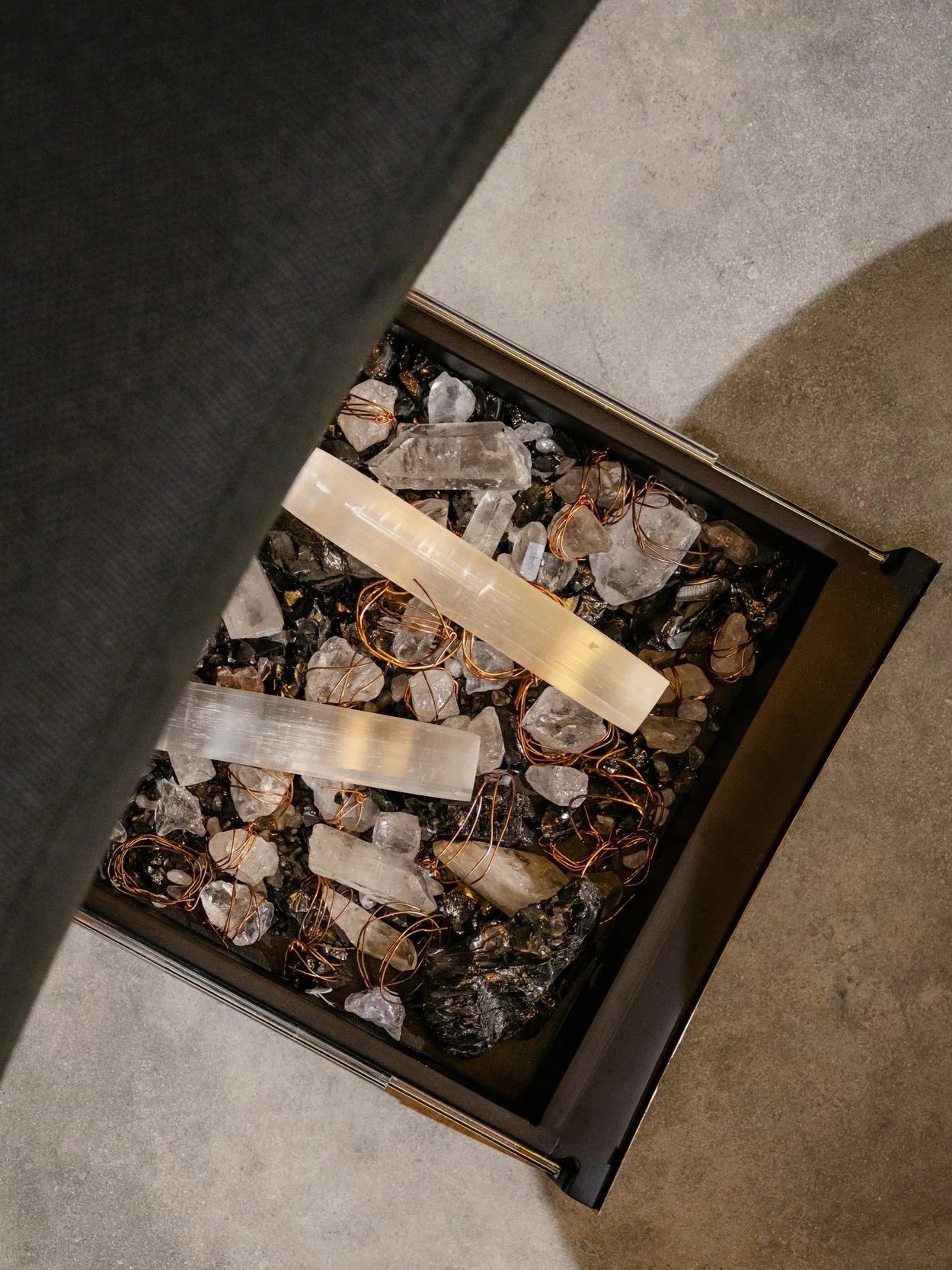 A floor-level decorative piece featuring a black rectangular container filled with clear quartz crystals, black stones, and copper wire accents, positioned next to a textured gray floor and a dark fabric surface.