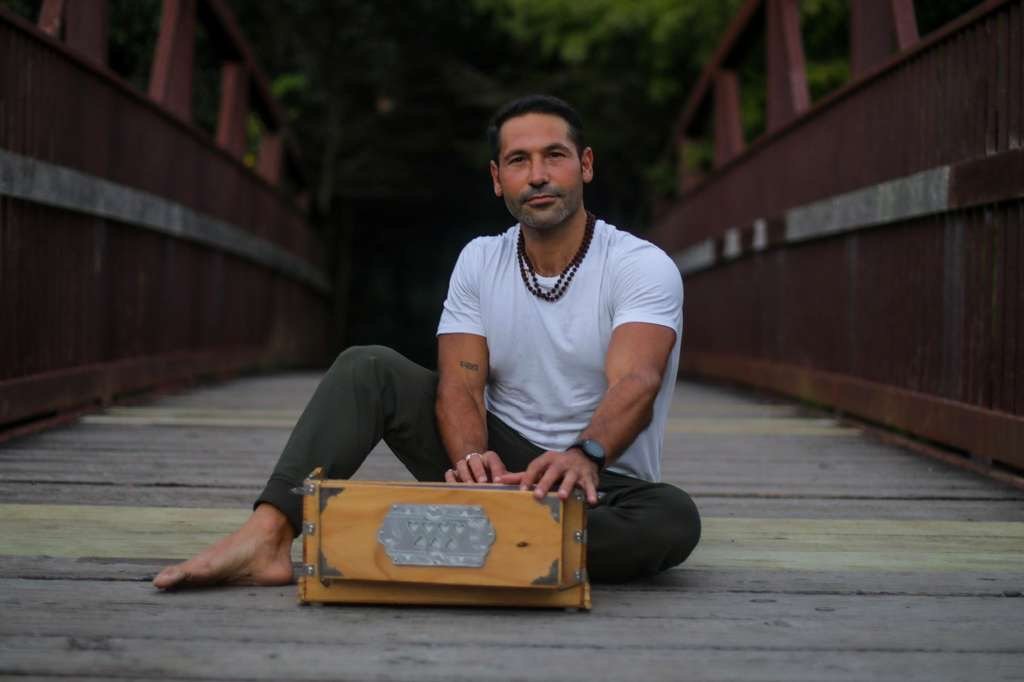 A man sits on a wooden bridge with greenery in the background, holding a wooden box with metal accents, wearing a white t-shirt, dark pants, a beaded necklace, a wristwatch, and no shoes.