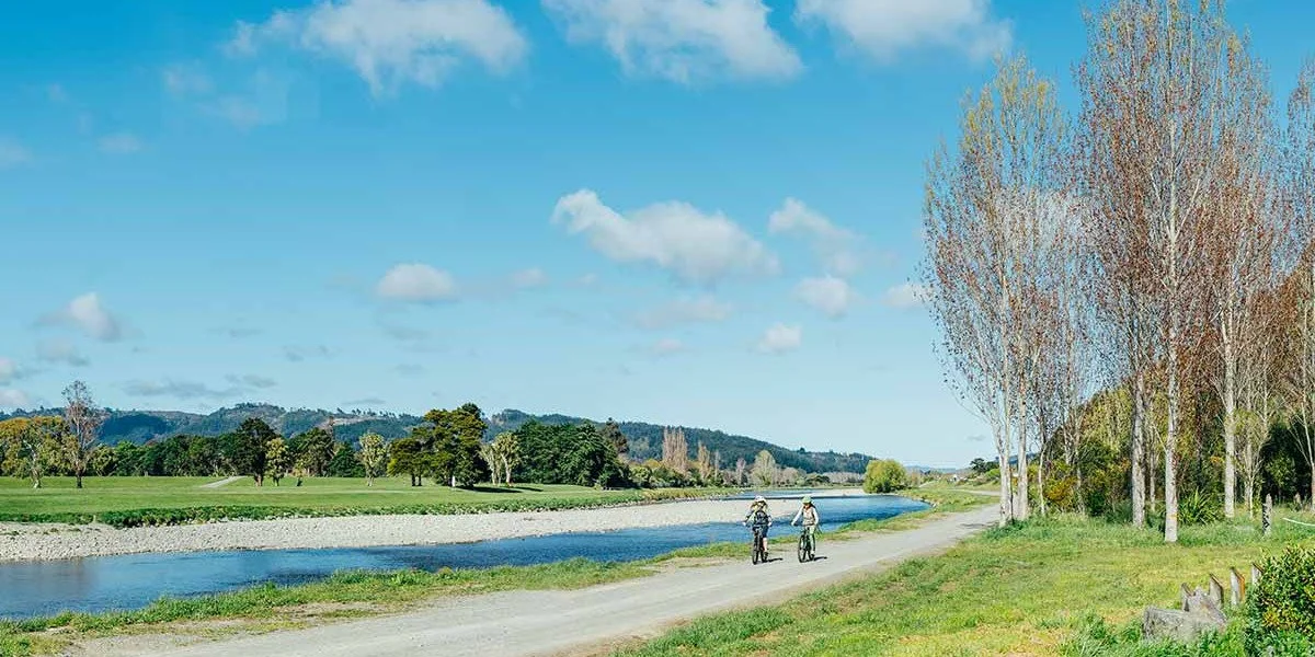 Two people riding bicycles on the Hutt River Trail next to a river, with trees and rolling hills in the background under a partly cloudy sky.
