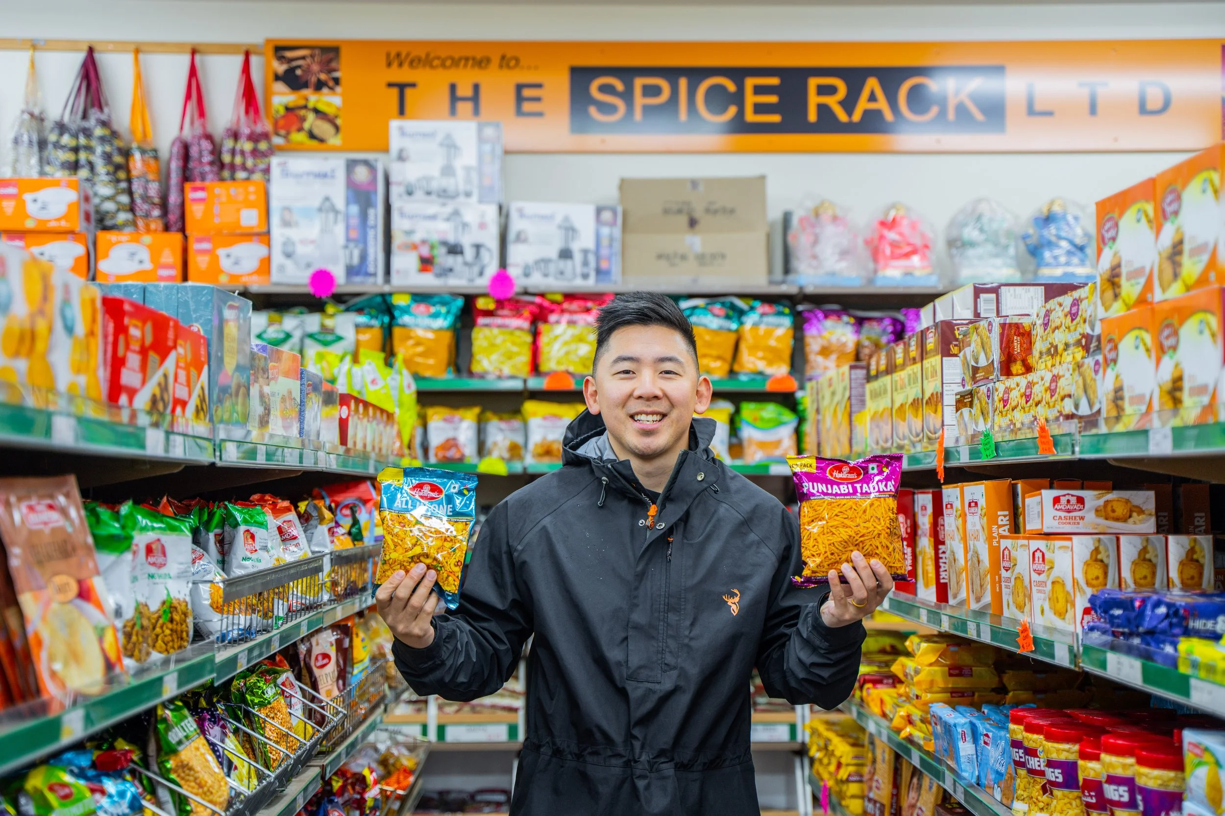 A smiling man in a black jacket shopping for snacks in an aisle of The Spice Rack in Petone, holding two packages of snack food.