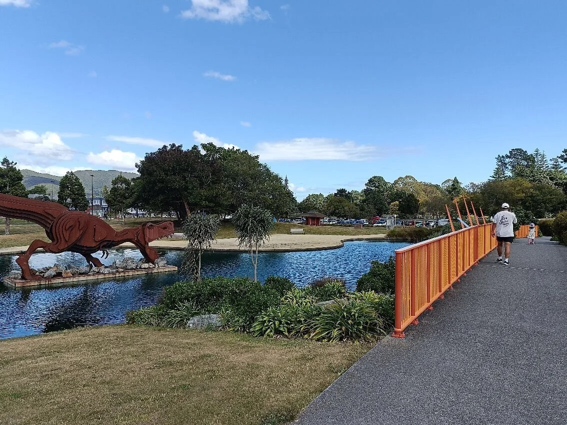 Avalon Park with a small pond, a large metal dinosaur sculpture, trees, a walking path with an orange railing, and a person walking away from the camera on a clear day.