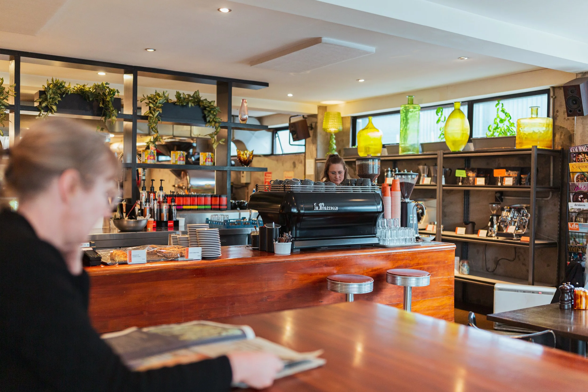 Interior of Revive Cafe in Petone, with a barista working behind the counter. Decor includes large green glass bottles and potted plants on shelves, with sunlight coming through large windows.