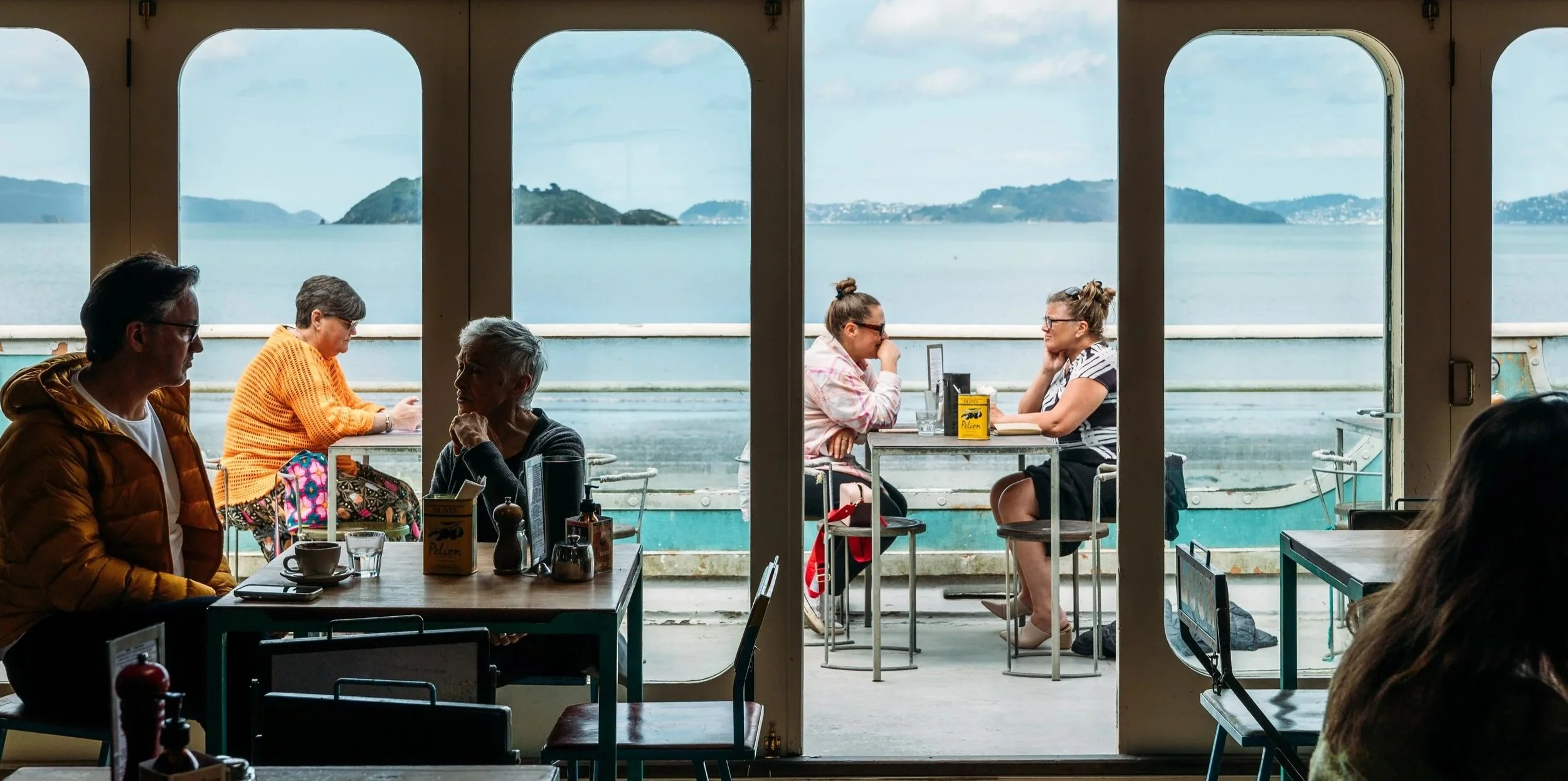 A view from inside Seashore Cabaret in Petone, showing several groups of people sitting at tables, with an outdoor scene of a body of water and islands through large windows behind them.