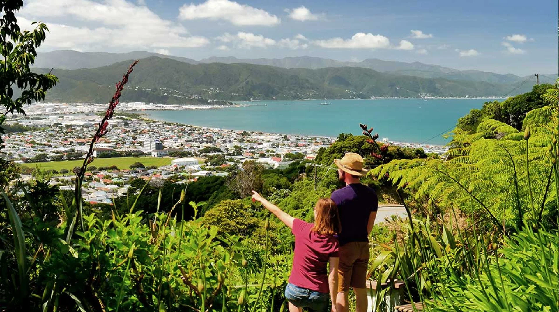Two people are standing on a trail in Percy Scenic Reserve, pointing towards a cityscape with a large body of water and mountains in the background, surrounded by lush green vegetation.
