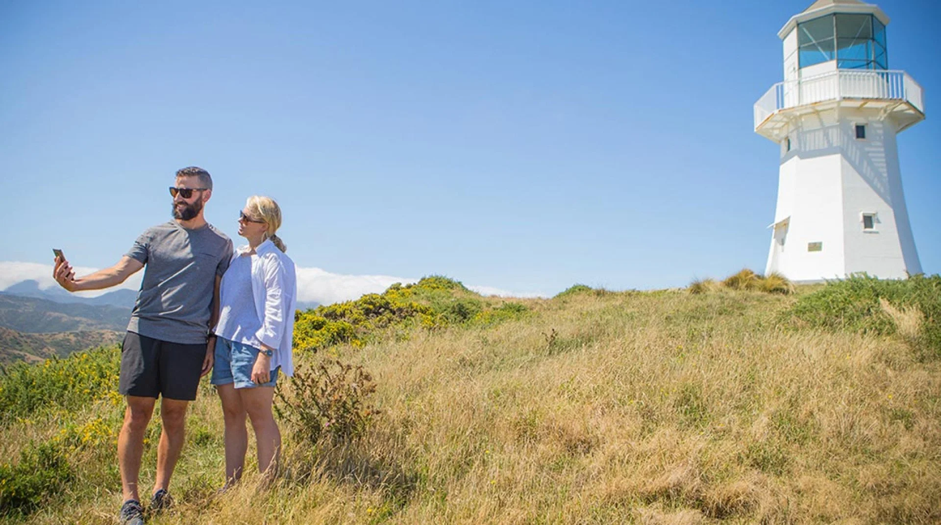 Two people, a man and a woman, taking a selfie outdoors near a white lighthouse on a grassy hill under a clear blue sky.