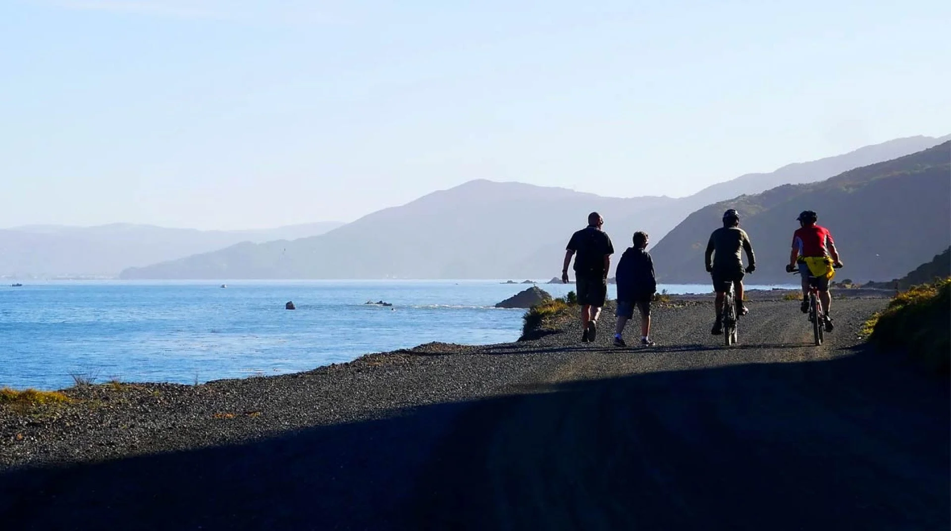 Four people, two walking and two biking, on a gravel pathway along a coastal area with mountains in the distance and the ocean on the left side of the image.