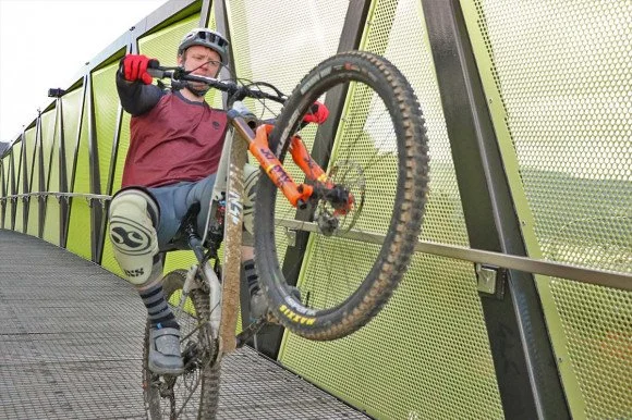 At Wainuiomata Mountain Bike Park, a person wearing a helmet and gloves is riding a mountain bike across a green metal bridge, raising the front wheel in a stunt.