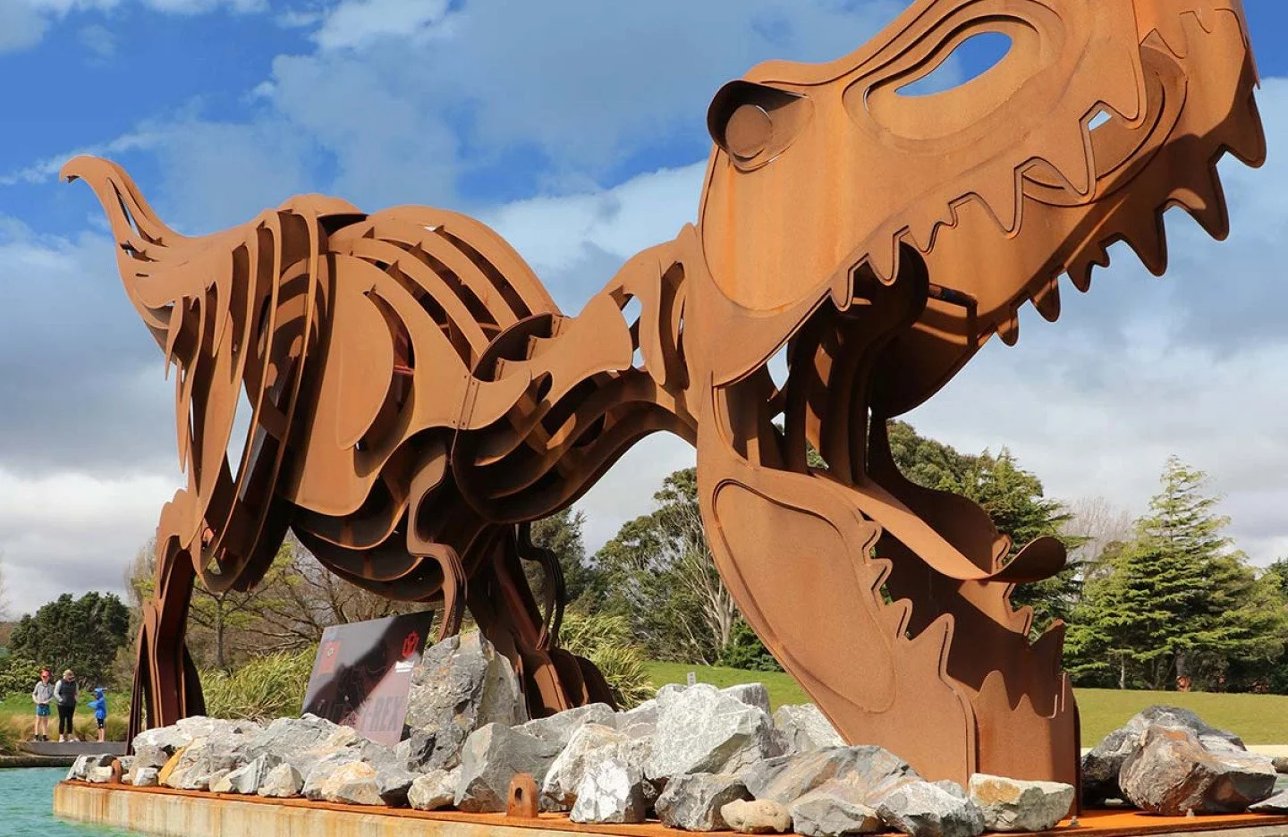 Located in Avalon Park is a large rust-coloured metal sculpture of a dinosaur skeleton with rocks at its base, outdoors with trees and cloudy sky in the background.