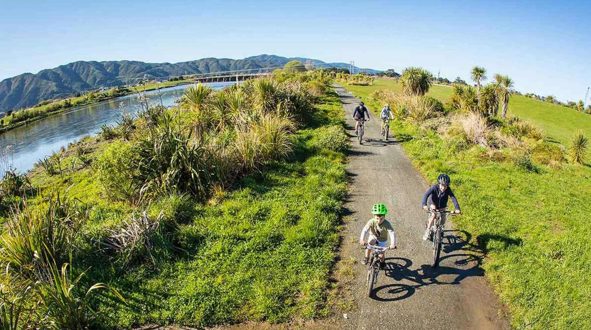 A group of children biking on the Hutt River Trail alongside a body of water with lush green vegetation and rolling hills in the background under a clear blue sky.