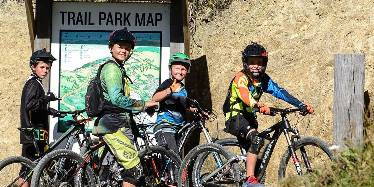 Four children in cycling gear with helmets on mountain bikes standing in front of a trail park map sign for Wainuiomata Mountain Bike Park