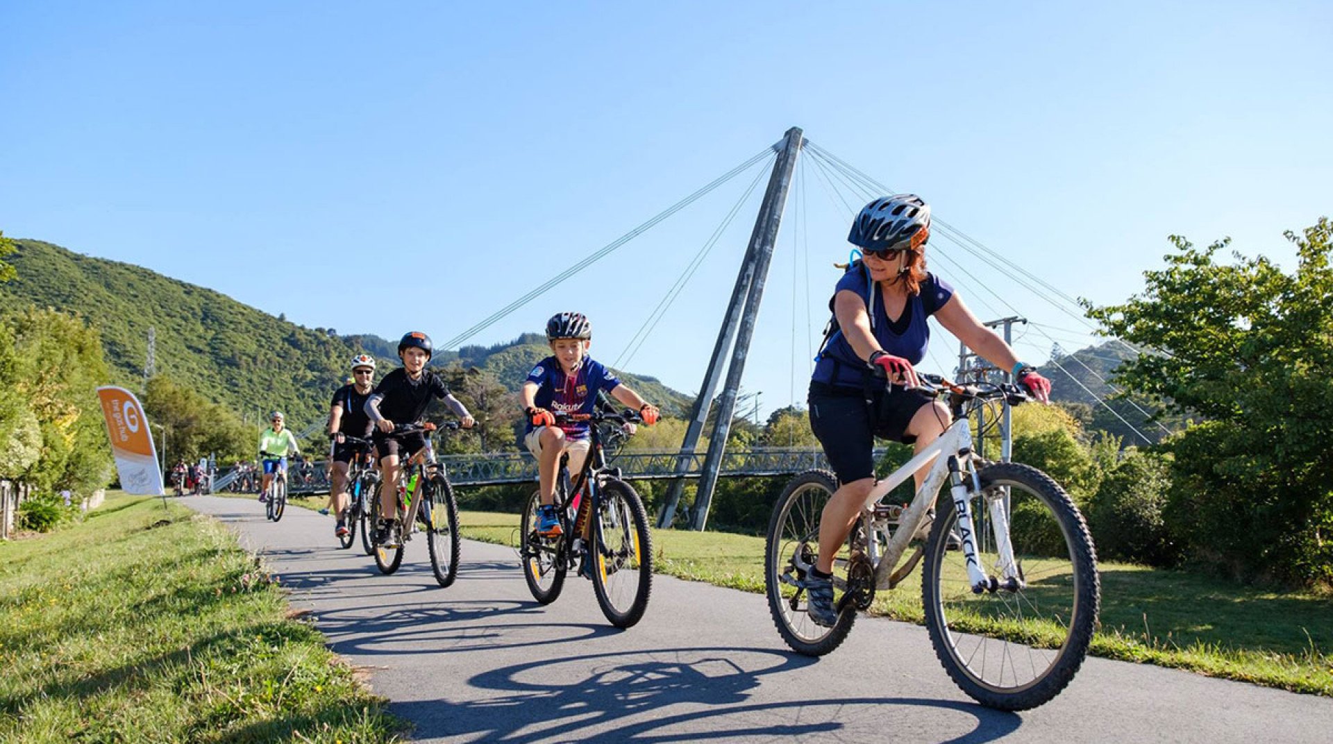 A group of children and an adult riding bikes on the Hutt River Trail in a scenic outdoor area with green hills, trees, a bridge, and power lines in the background on a sunny day.