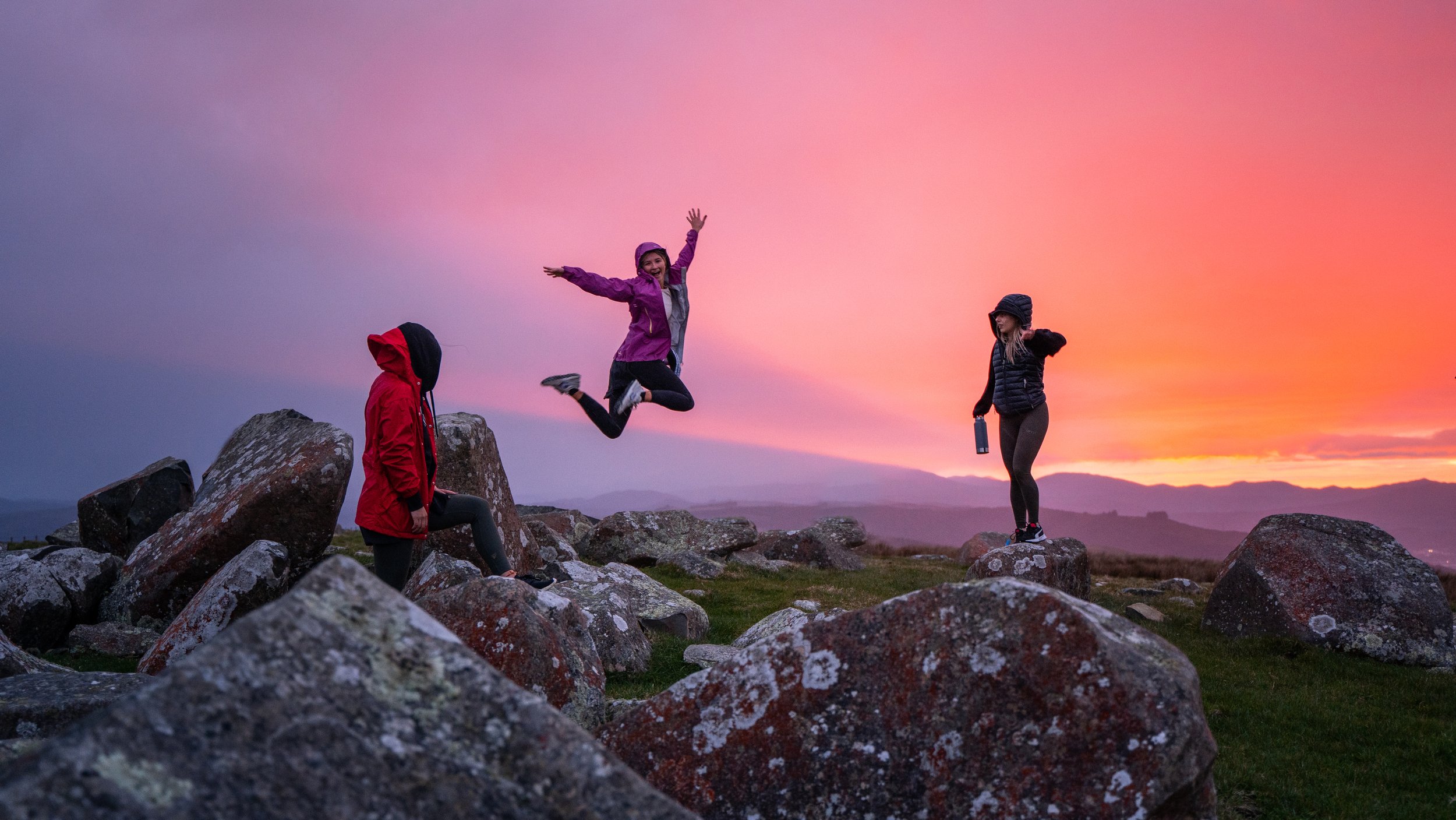 Three women are on Boulder Hill in Belmont Regional Park during sunset, one jumping with arms raised, others standing, dressed in outdoor gear.