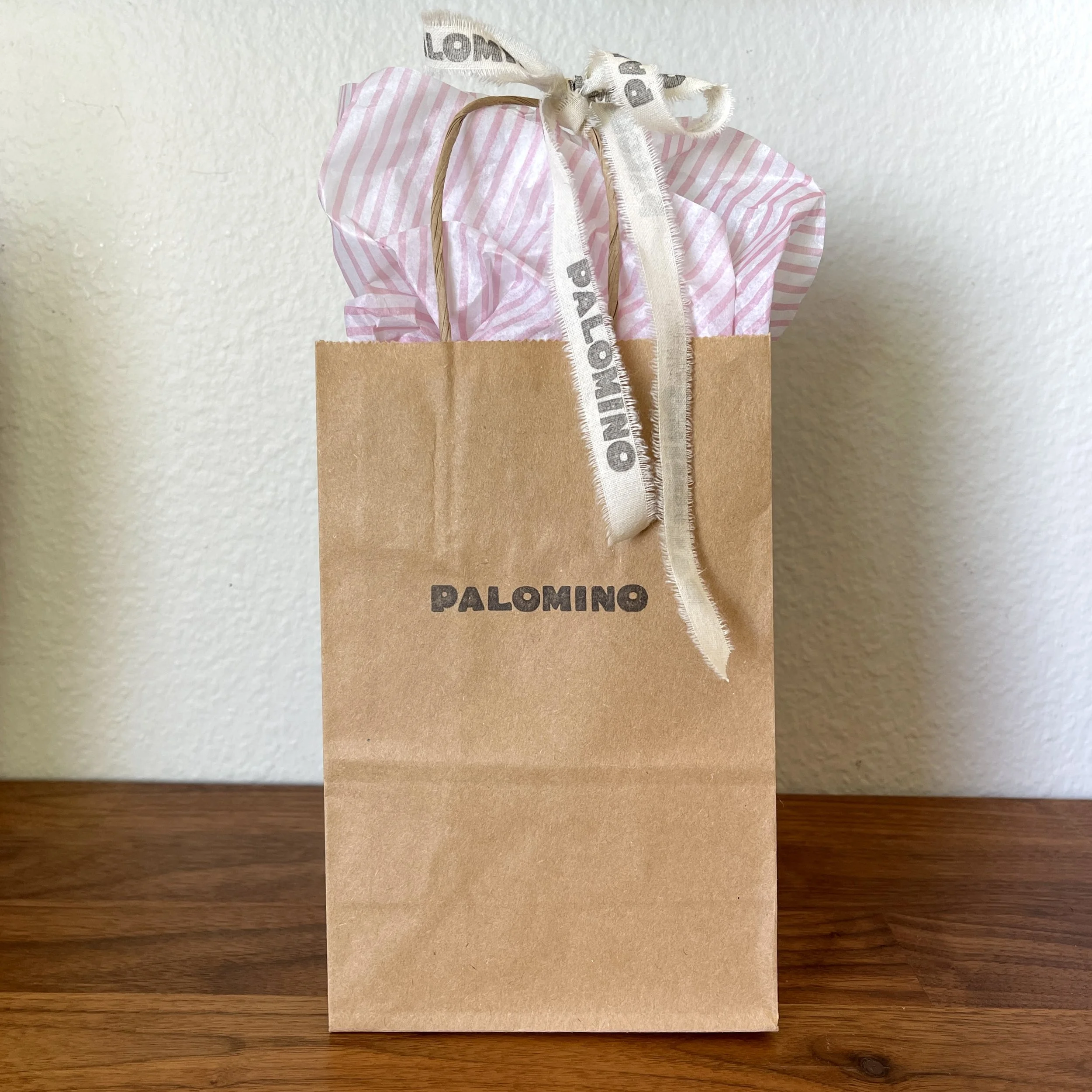 A paper gift bag on a wooden surface with pink and white striped tissue paper inside, tied with a white ribbon that has black text.