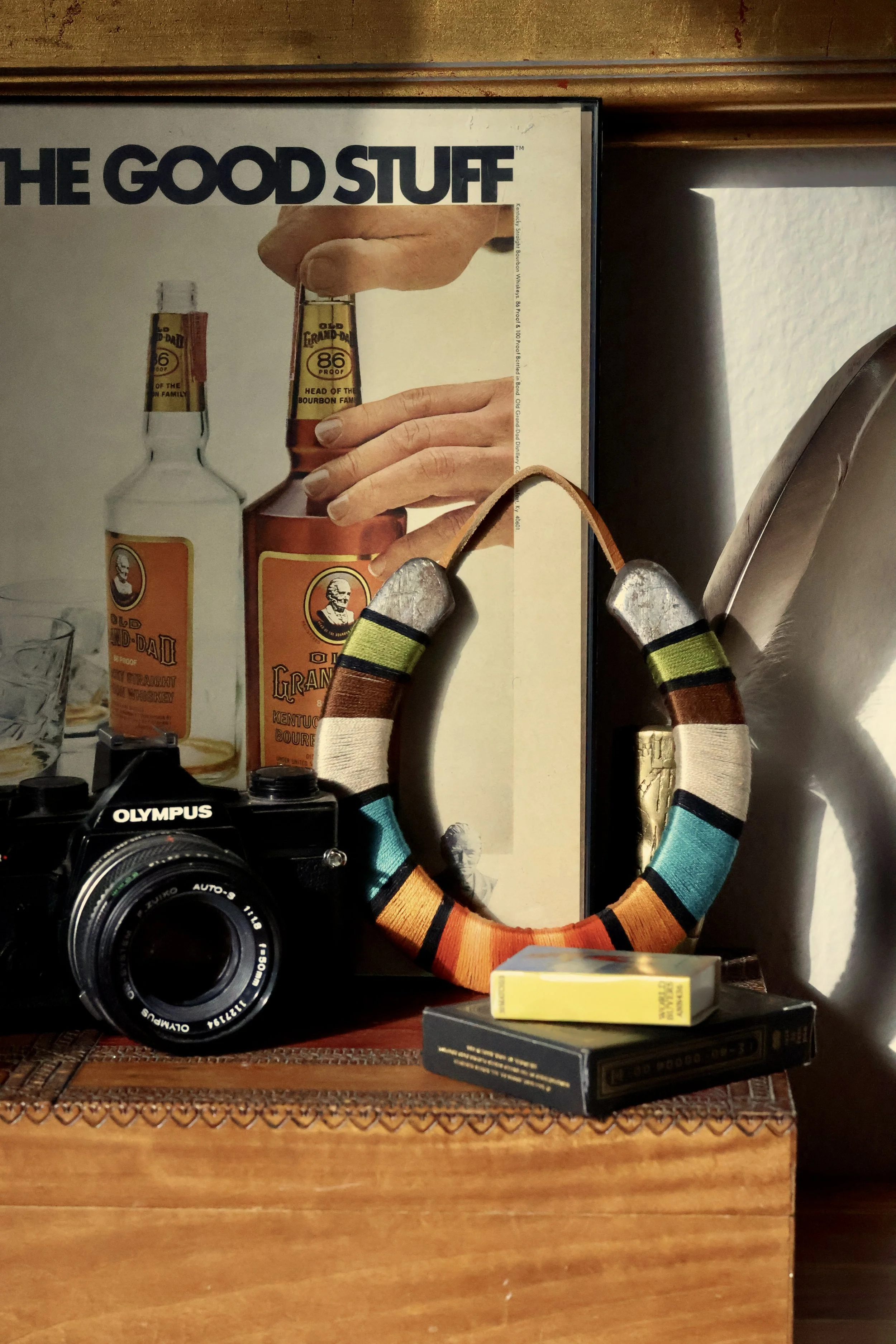 A framed vintage advertisement for Old Grand-Dad Kentucky Bourbon, depicting a hand holding a bourbon bottle. The background shows part of a wooden surface, a mirror, and some small objects, including a camera and a box.