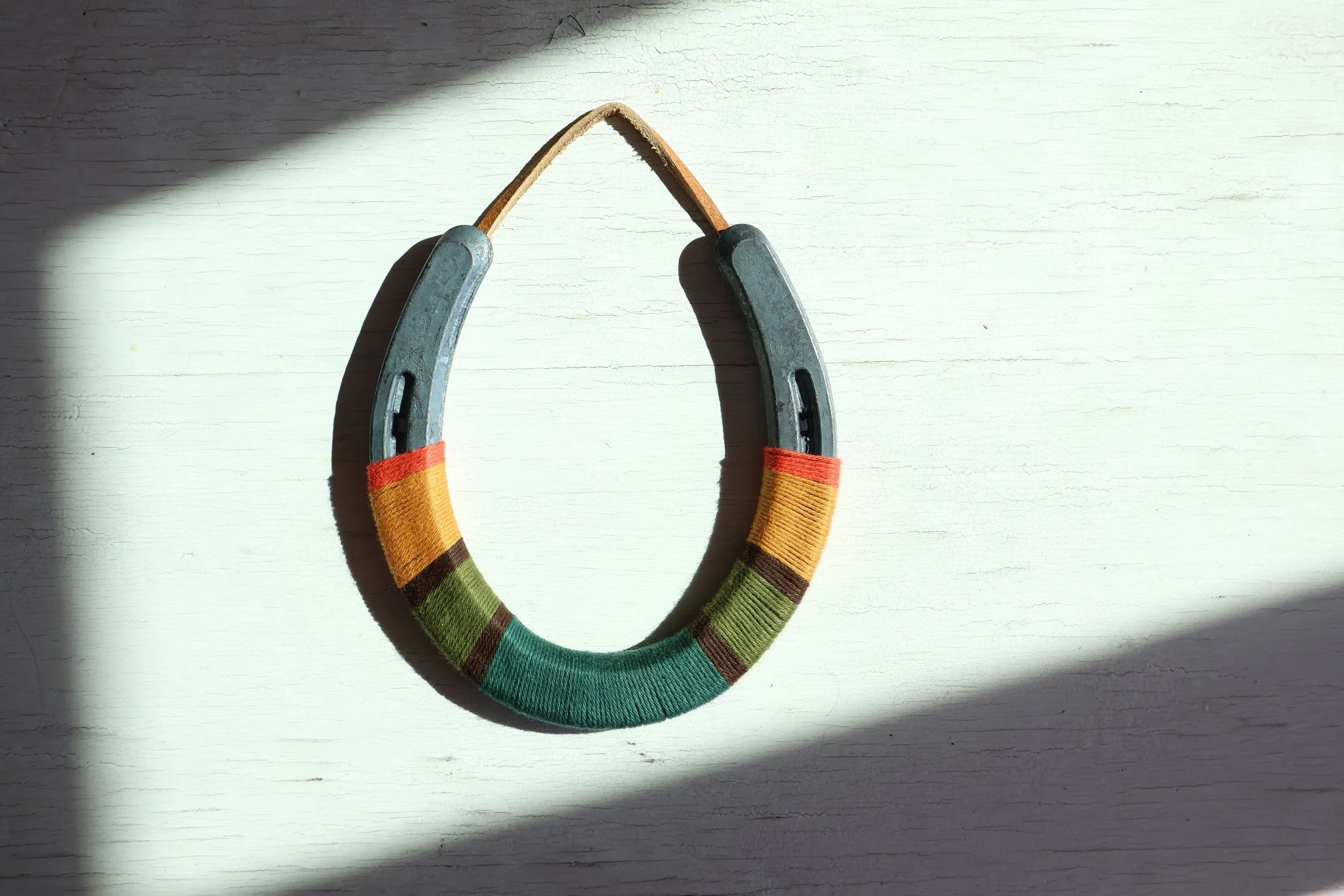 A horseshoe with a colorful, striped fabric covering on the bottom half, placed on a white wooden surface with light and shadow.