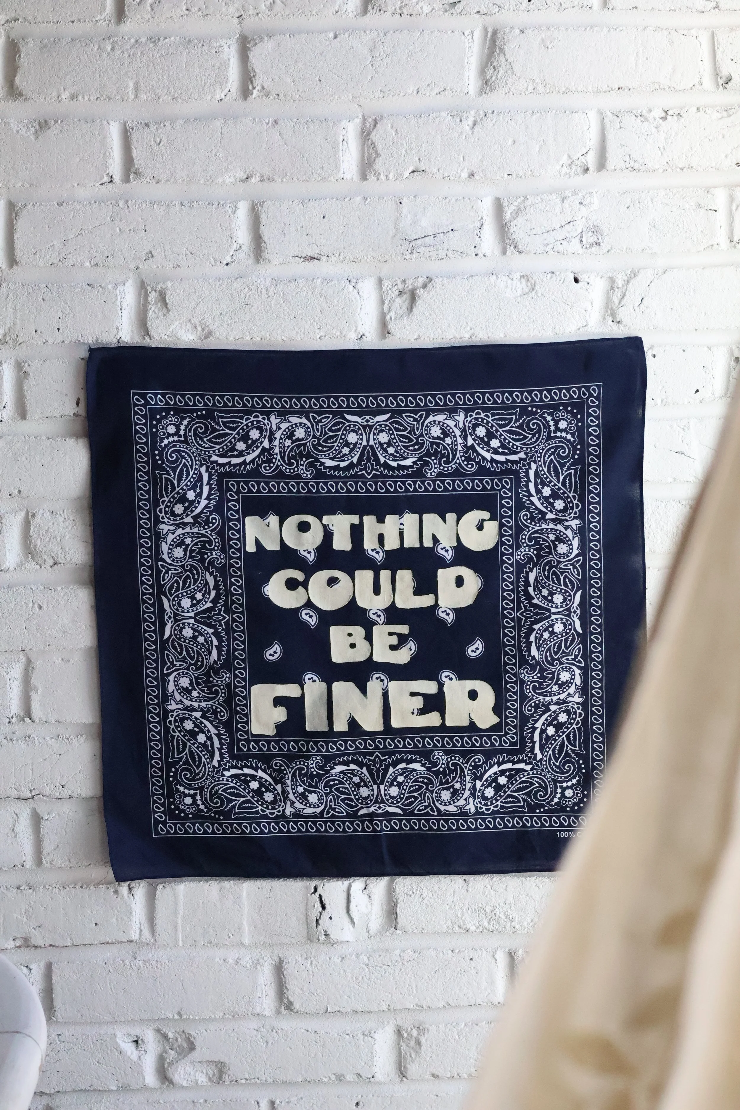 A blue bandana with white decorative patterns and the phrase 'Nothing could be finer' printed on it, hanging on a white brick wall.