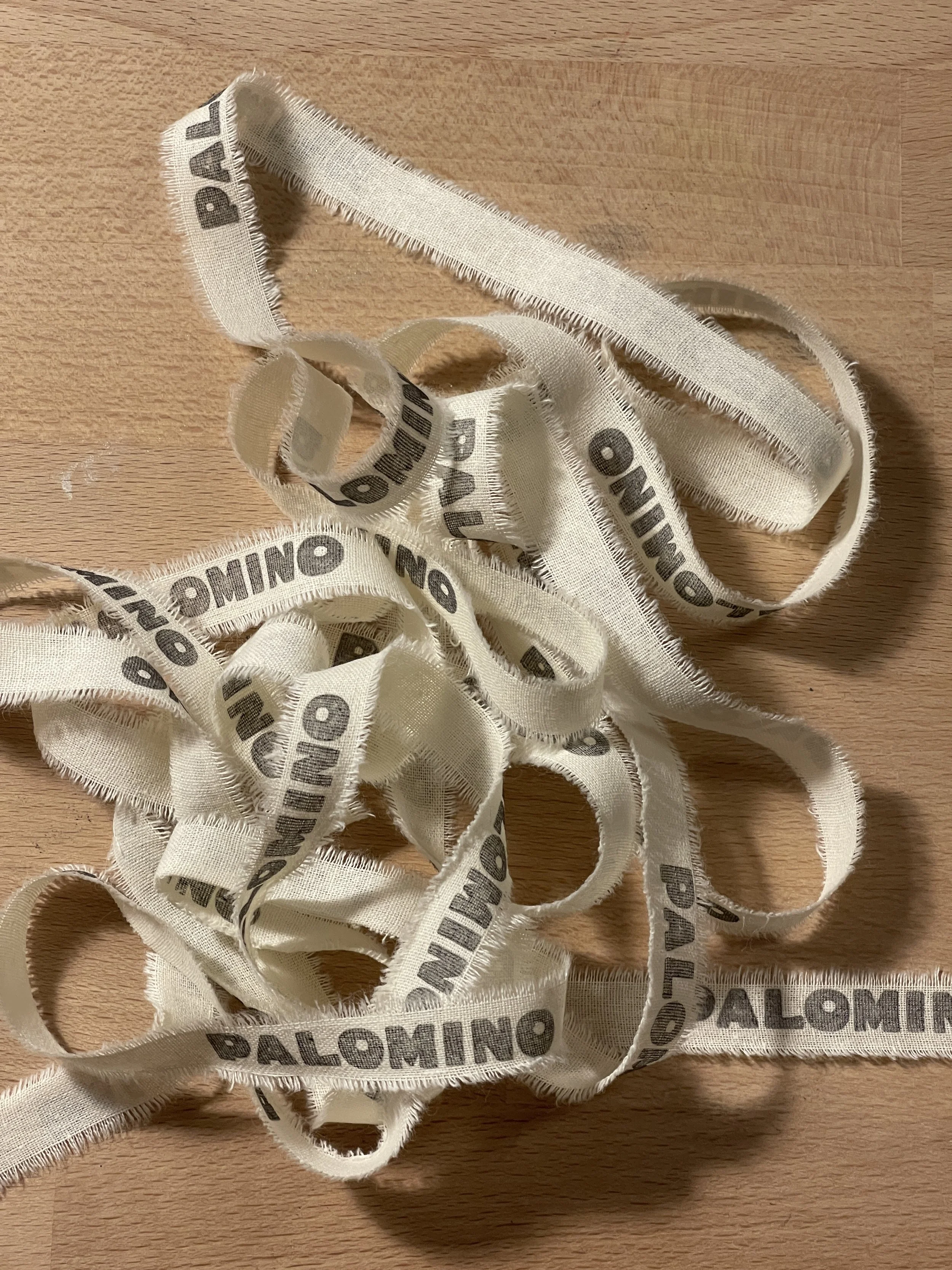 Crumpled white fabric ribbons with the word 'PALOMINO' printed repeatedly in black, laid out on a wooden surface.