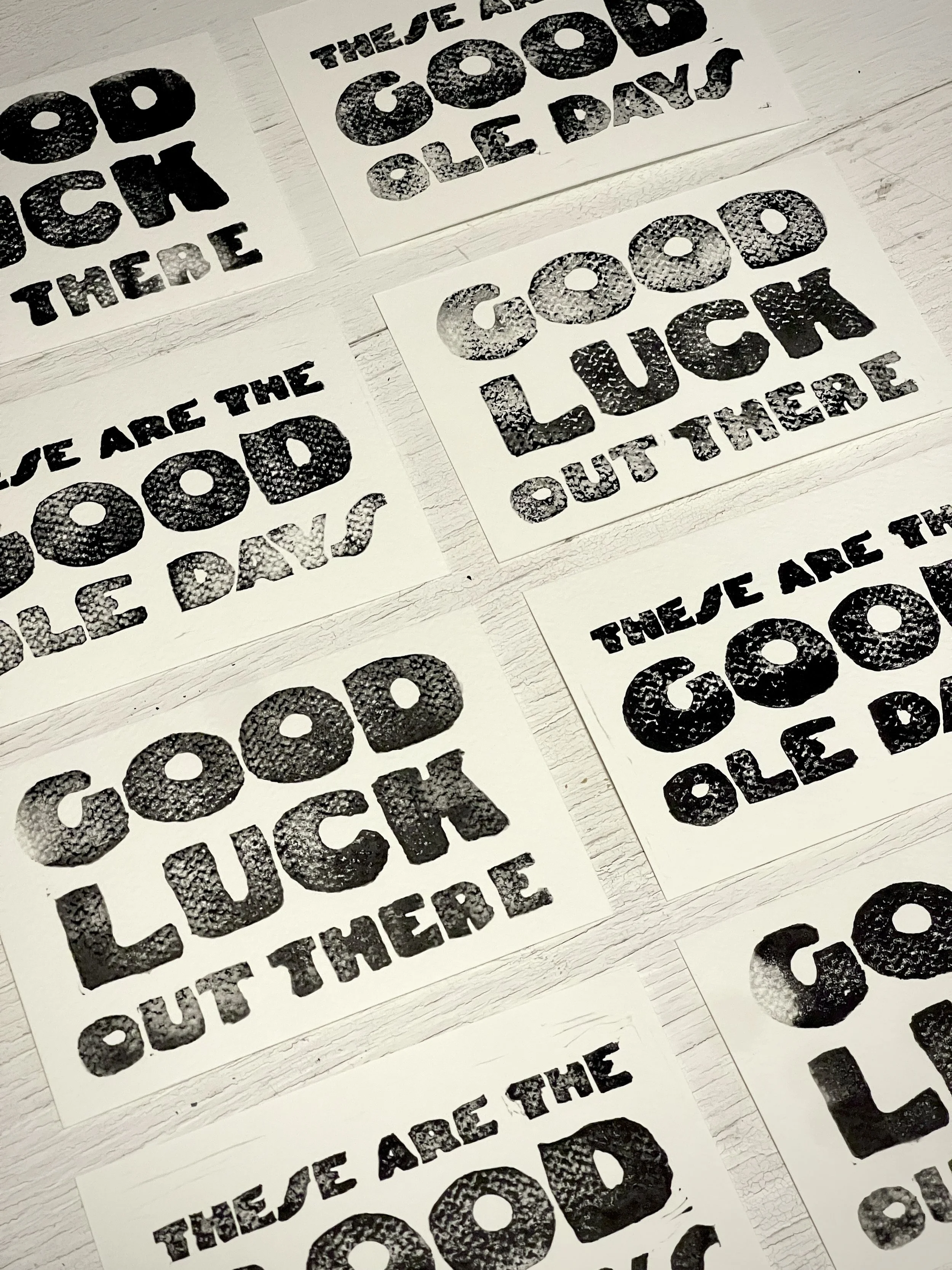 Multiple sheets of paper with black and white stencil-style text and images, including the words 'OCD', 'THE LITTLE AÑO', 'LOVE', and 'OUT THERE', arranged on a white wooden surface.