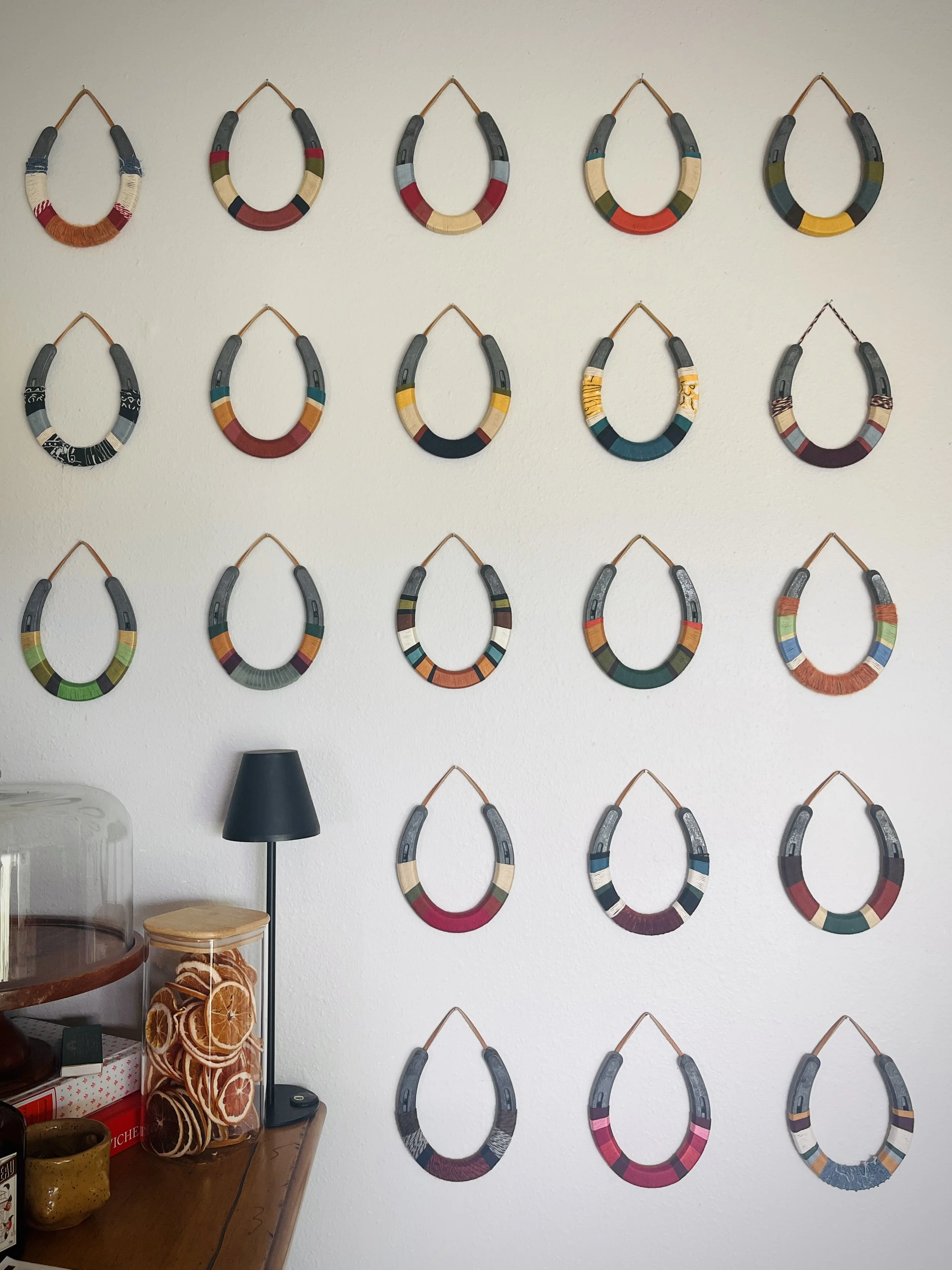 Colorful woven jewelry necklaces hanging on a white wall in a grid pattern, with a table, lamp, and decorative orange slices in the foreground.