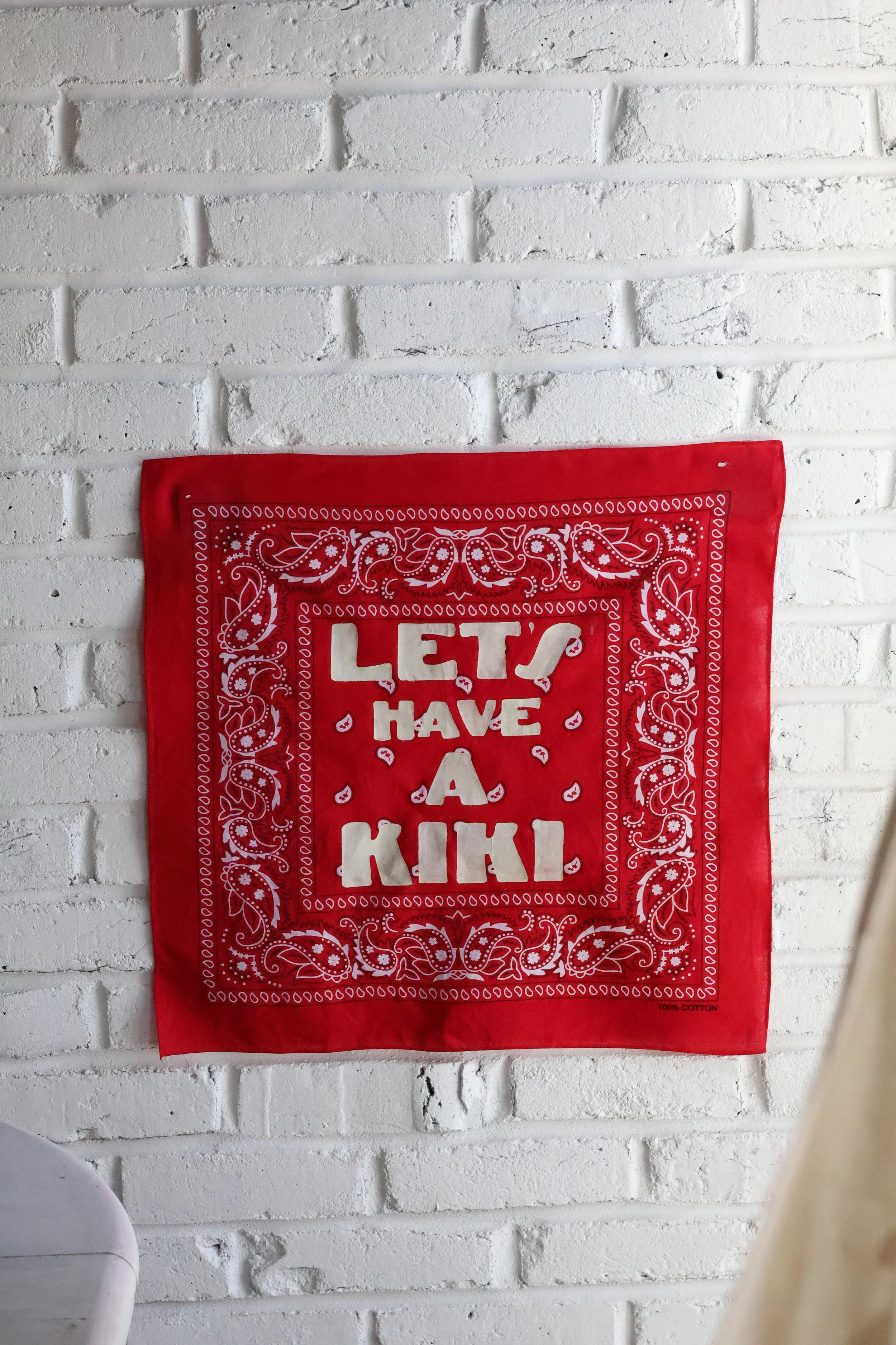 A red bandana with white paisley patterns and the text "Let's Have a Kiki" is hanging on a white painted brick wall.
