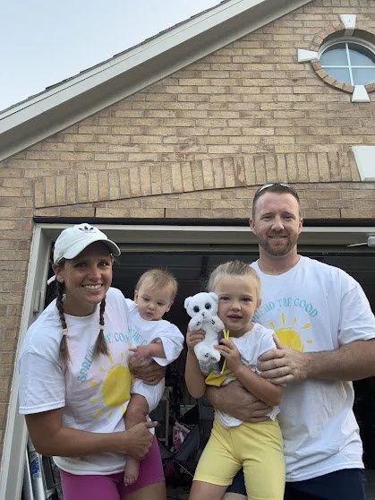 A smiling family of four stands outside their house, holding a stuffed polar bear and a young girl, all wearing casual summer clothes with a matching theme and white T-shirts with a yellow sun design.