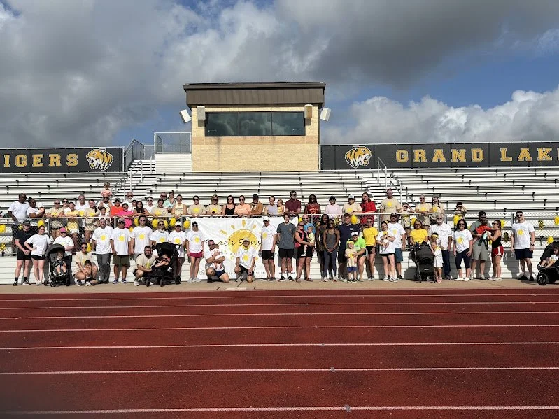Group of people gathered on stadium bleachers, holding a banner and wearing yellow and white t-shirts, in front of a track at Grand Lake Tigers stadium on a cloudy day.