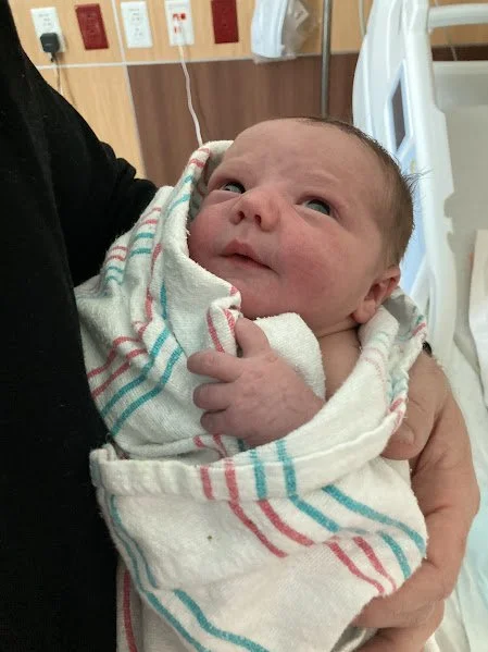 Newborn baby wrapped in a hospital blanket held by a person, in a hospital room with medical equipment.