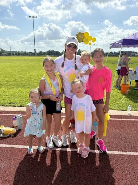 Group of six women and children smiling on a track field, wearing colorful athletic clothes, with balloons and tents in the background.