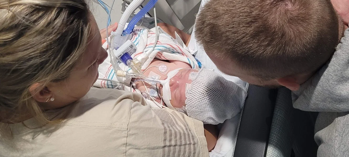 A woman and a man are sitting beside a hospital bed with a newborn baby in it, connected to medical tubes and monitors.