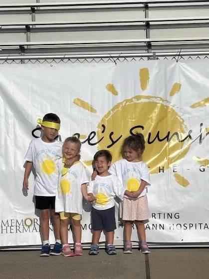Four children standing in front of a white banner with a yellow sun and the words 'Shae's Sunshine' and other partially visible text. The children are wearing white t-shirts with yellow sun designs and are smiling.