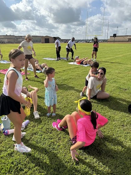 Children and adults on a football field participating in a fitness or sports activity day, some lying on the ground, others standing or sitting, with cloudy sky overhead.