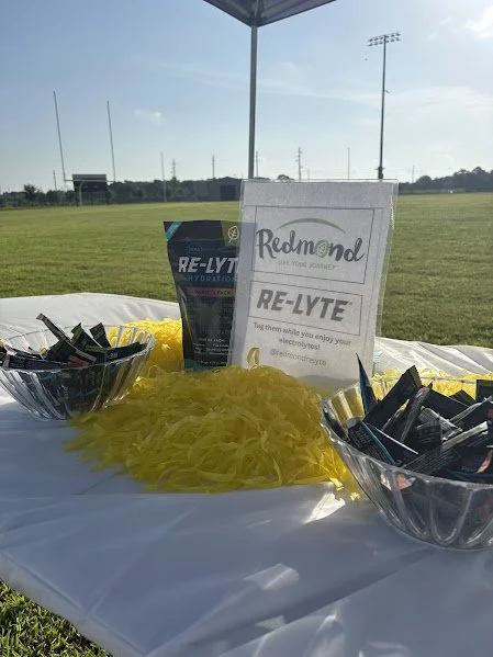 Table with bowls of individual Re-LYTE electrolyte packets, a yellow feather boa, and a sign for Redmond Re-LYTE at a sports field.