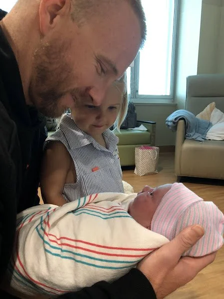 A man holding a newborn baby wrapped in a hospital blanket, with a young girl looking on with curiosity in a hospital room.