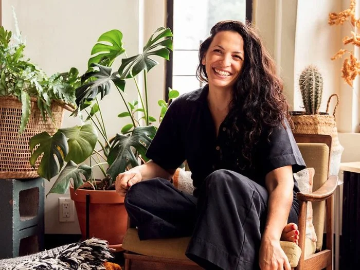 A woman with curly dark hair sitting on a yellow armchair in a room filled with green houseplants, smiling at the camera.