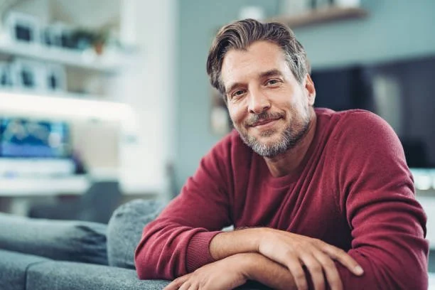 A middle-aged man with gray hair and a beard, smiling, sitting on a couch in a modern living room.