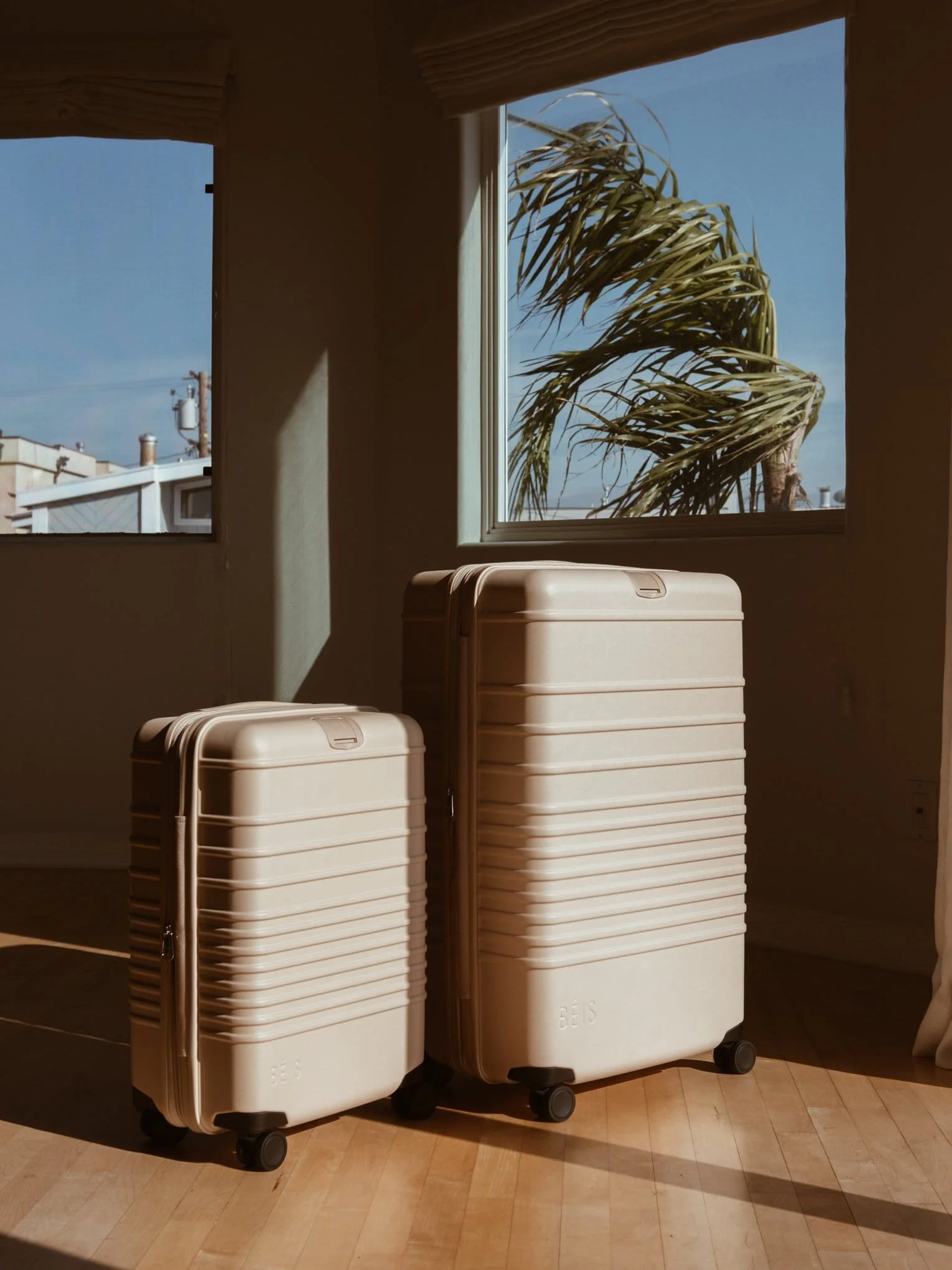 Two cream-colored rolling suitcases of different sizes placed on a wooden floor near a window with sunlight and a view of a palm tree outside.