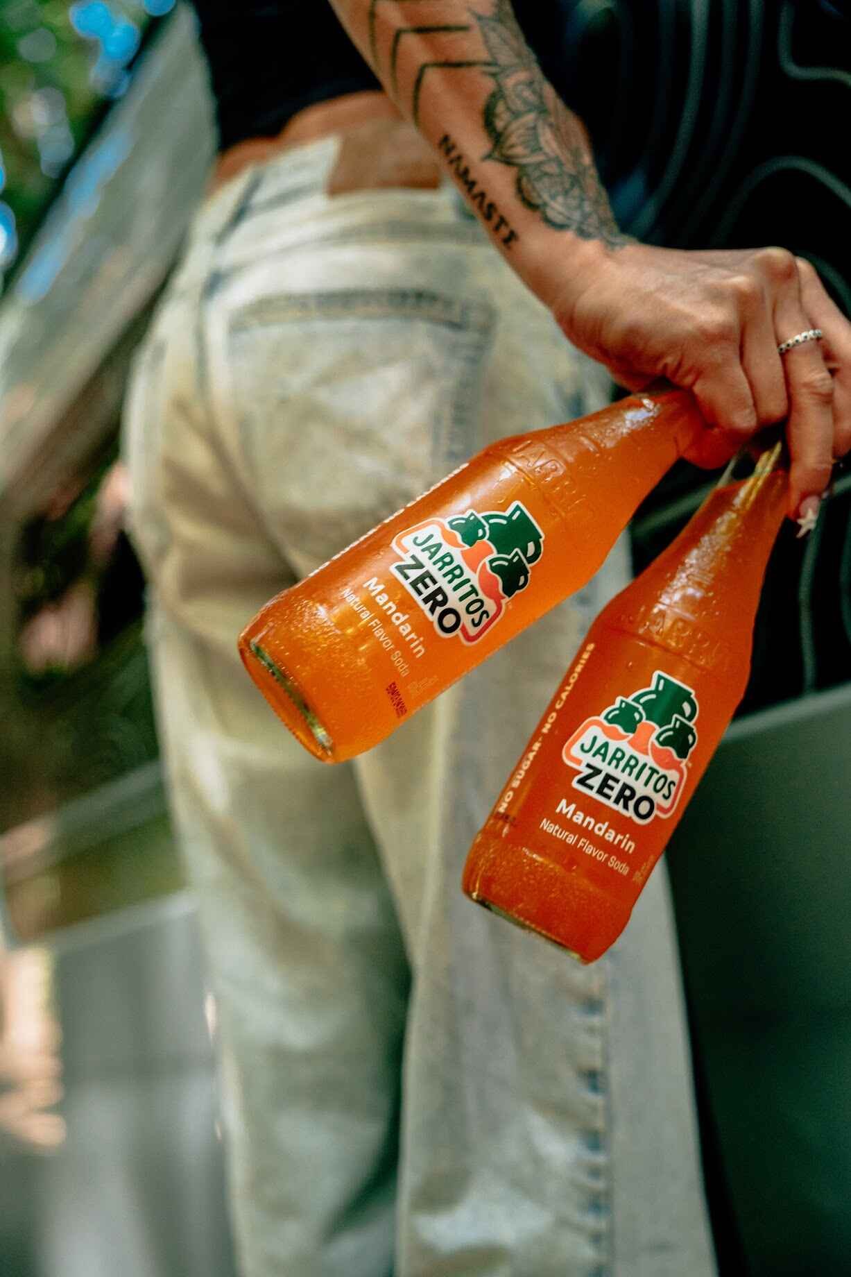 Person holding two bottles of Jarritos Zero Mandarin soda, with tattoos visible on their arm and wearing light-colored pants.