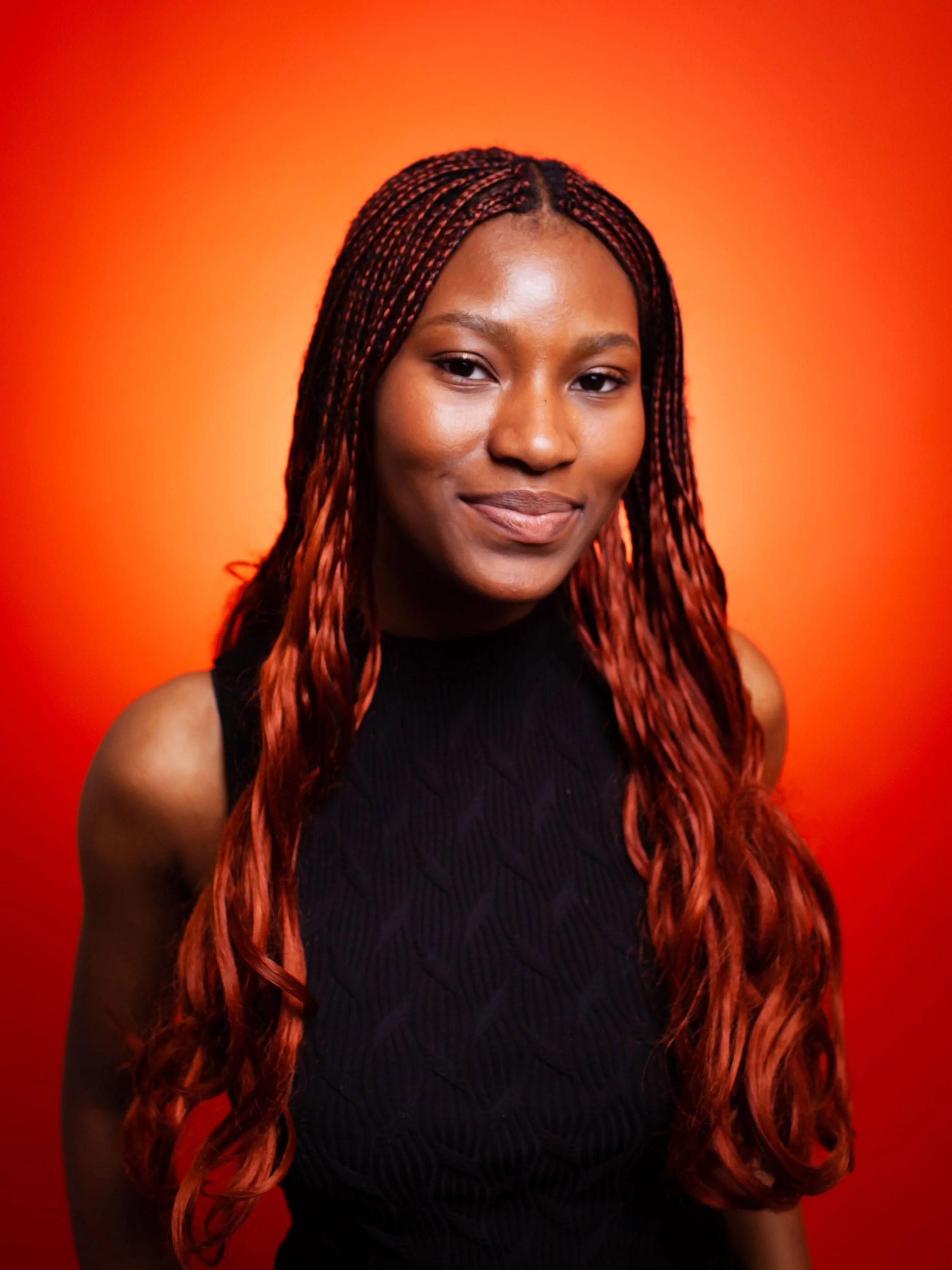 Portrait of a young woman with dark skin and long, wavy red hair, wearing a sleeveless black top, smiling gently against a vibrant orange background.