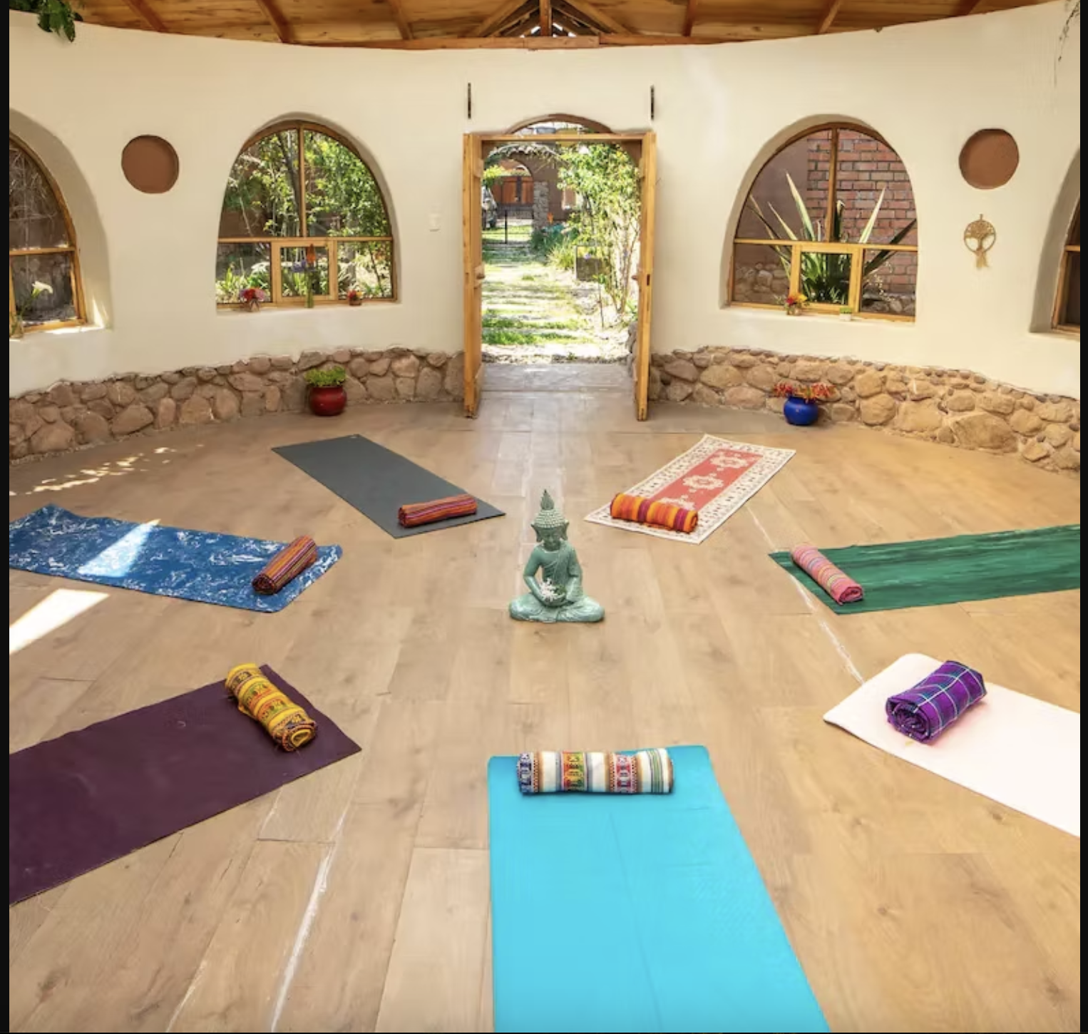 Yoga class setup with seven mats and rolled towels arranged in a circle around a small Buddha statue in a room with arched windows and a view of a garden outside.