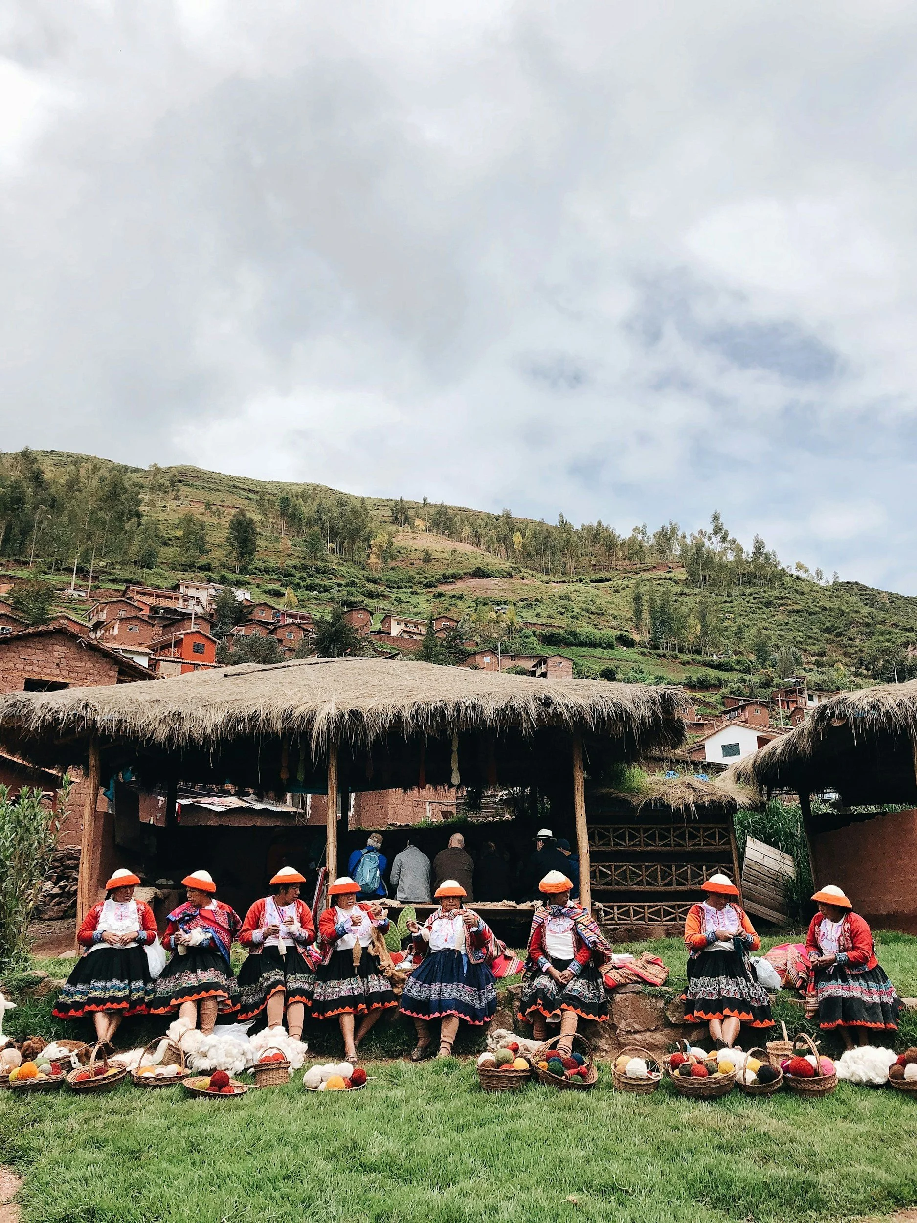 Women dressed in traditional clothing sitting on the grass with baskets of colorful yarn in front of a thatched-roof structure, with a hillside village in the background.