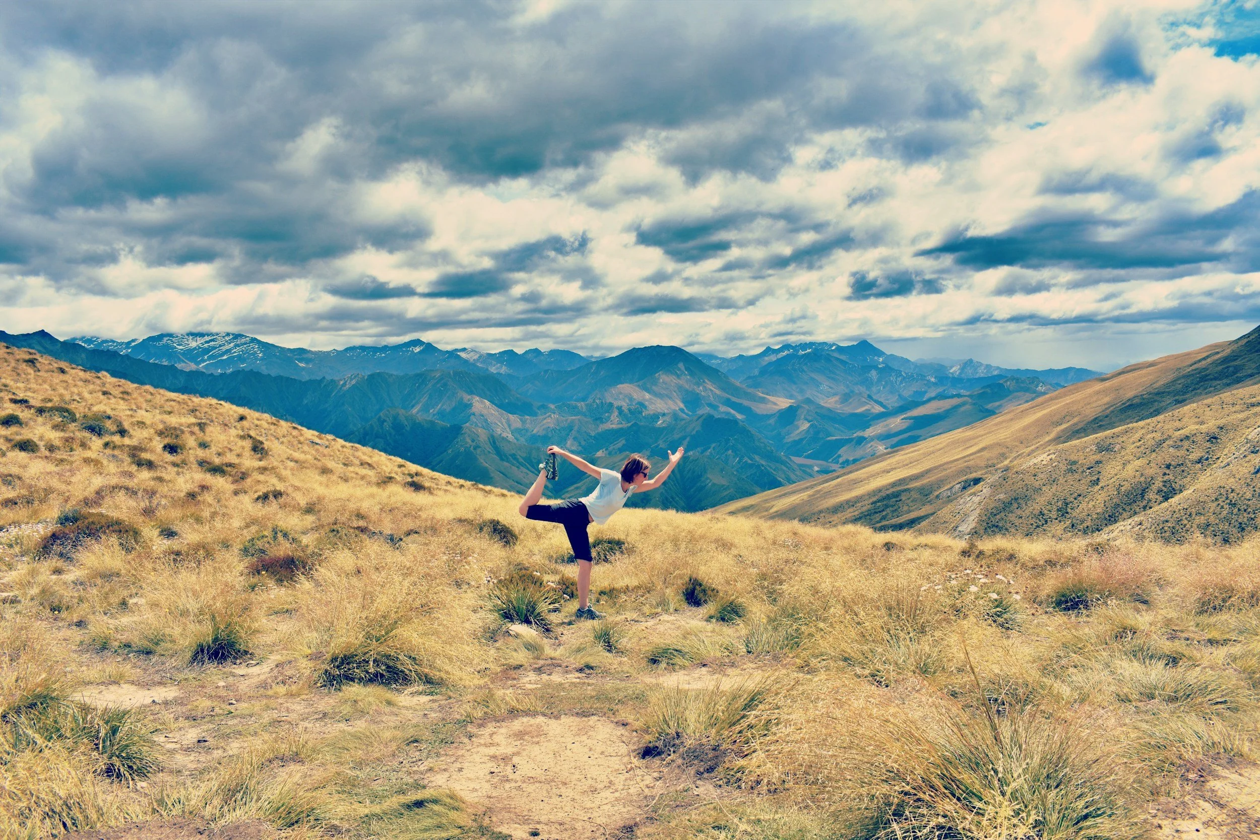 A person performing a yoga pose on a grassy hillside with mountains in the background, under a cloudy sky.