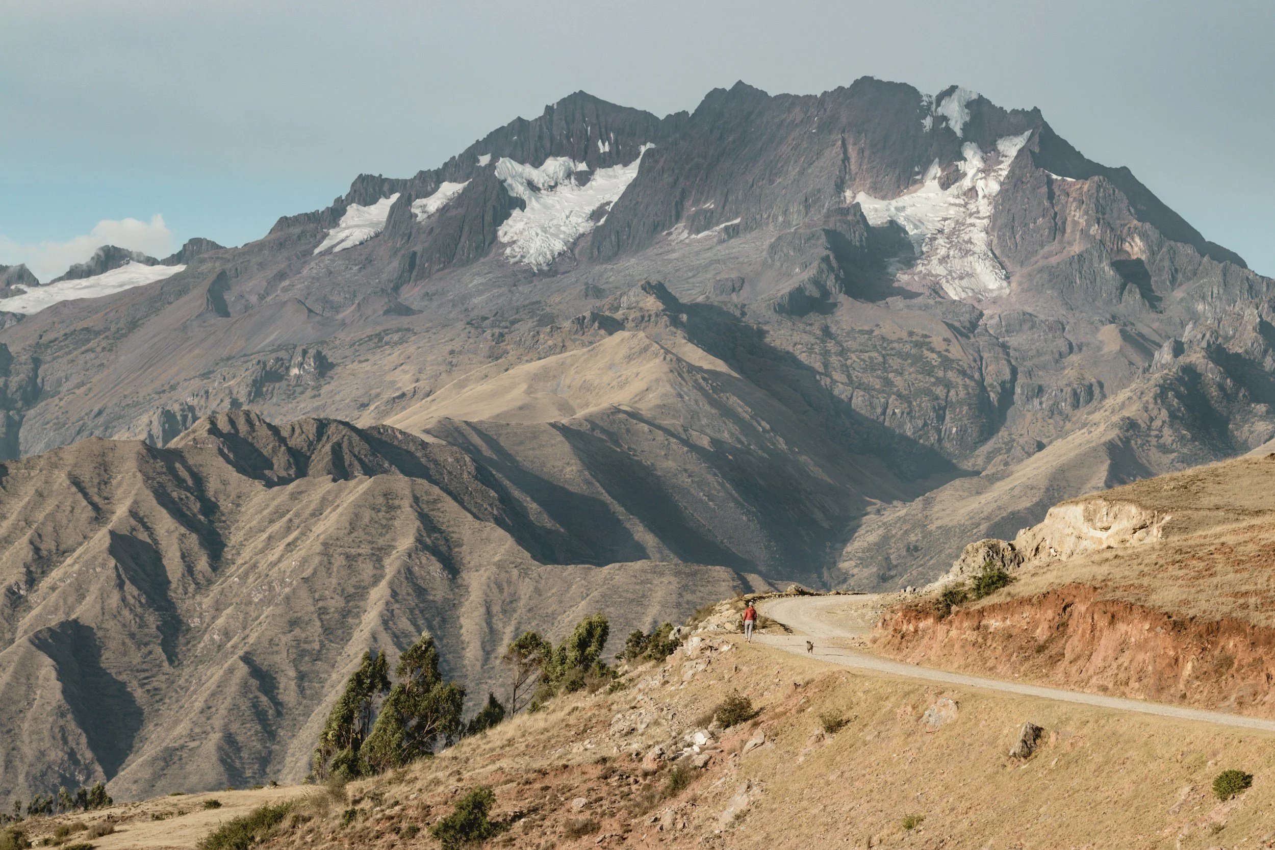 A mountainous landscape with rugged peaks, some with snow, and a winding dirt road with a person walking along it.