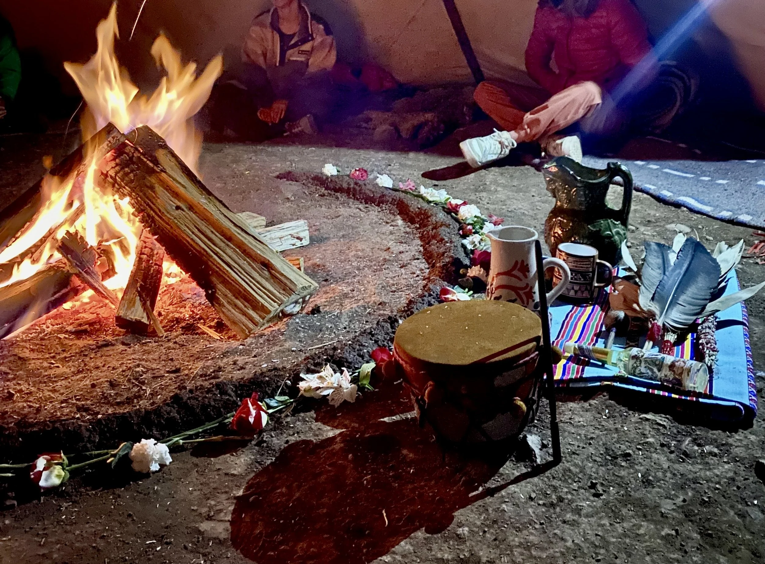 A campfire with logs burning inside a circle of flowers and small objects. Sitting on the ground are three children dressed warmly in jackets, with some personal items like a mug, a decorative metal pitcher, feathers, and other items placed on a colorful mat nearby.