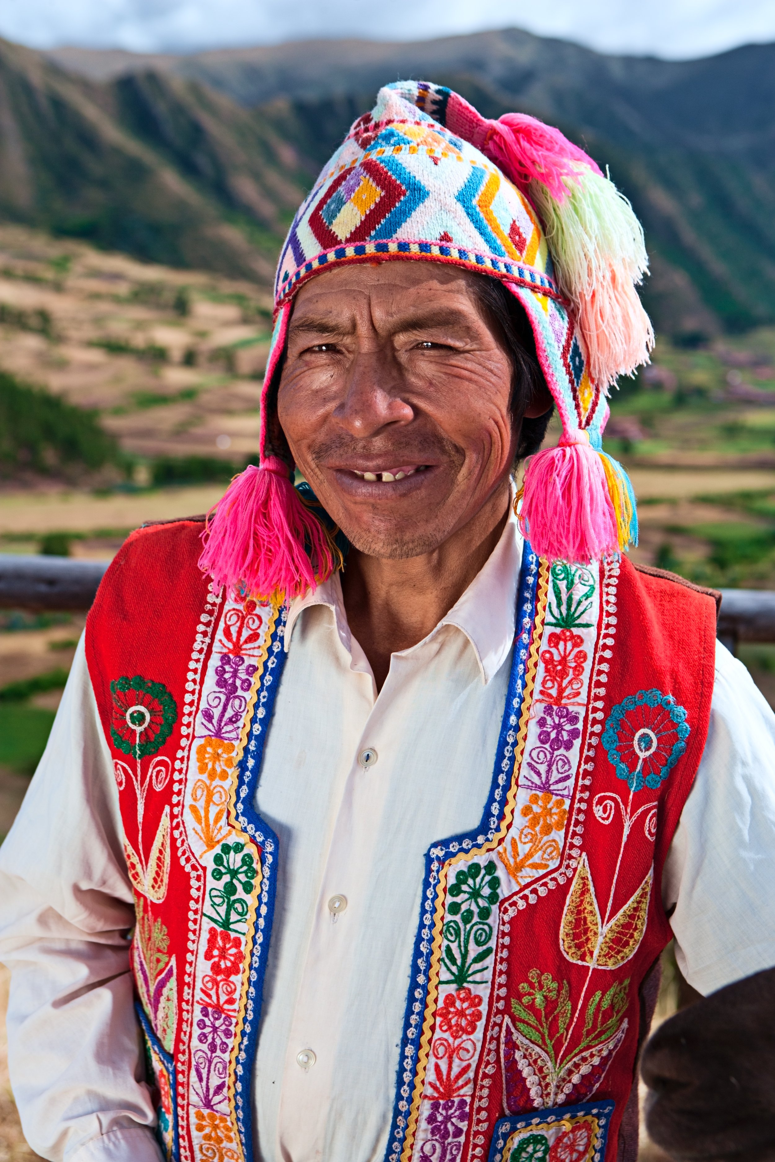 A man wearing traditional Mexican embroidered clothing and a colorful embroidered hat with pink tassels, smiling outdoors with a mountainous landscape in the background.