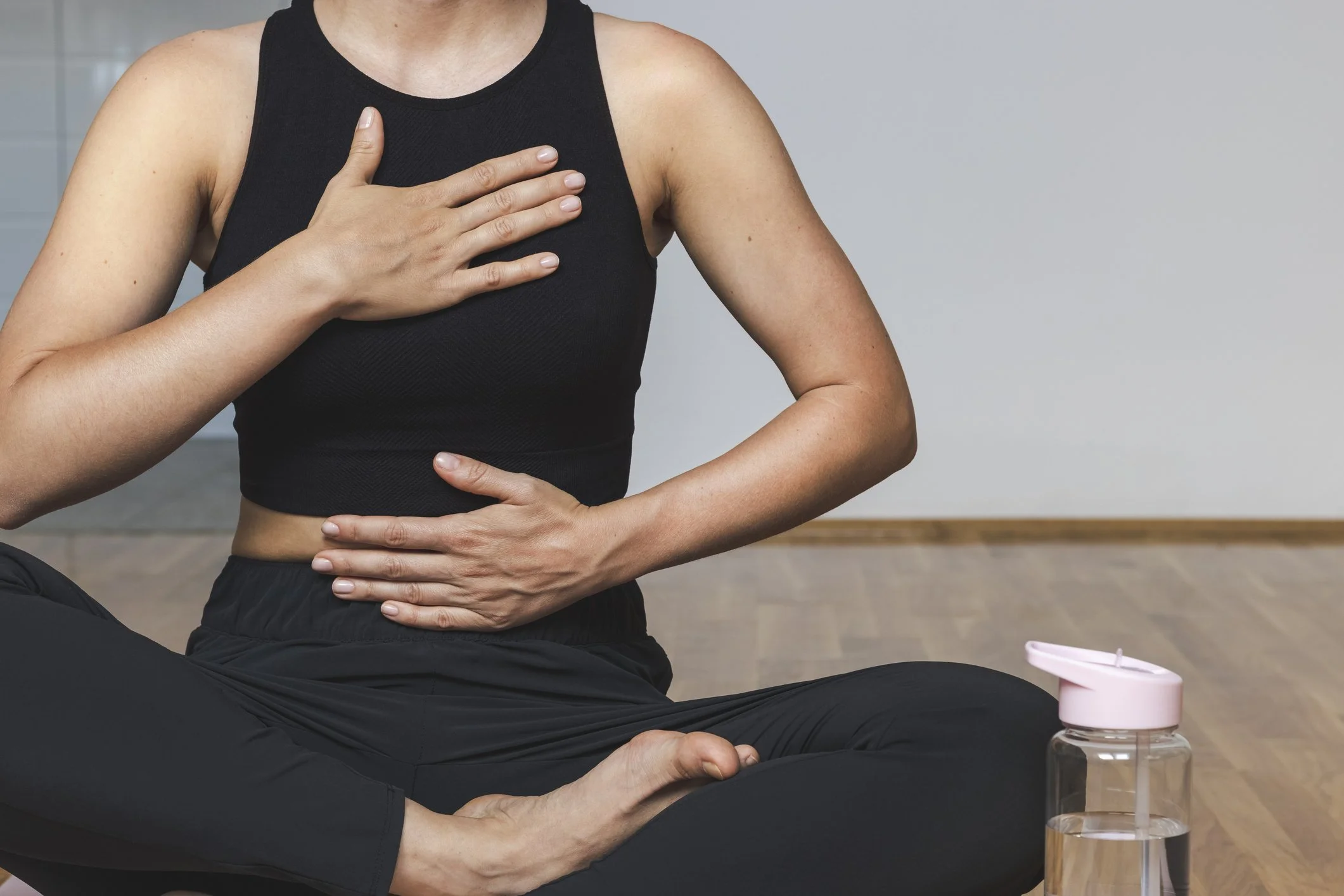 A person practicing yoga or meditation, sitting cross-legged on the floor, wearing a black tank top and black pants, with one hand on their chest and the other on their stomach, and a water bottle nearby.